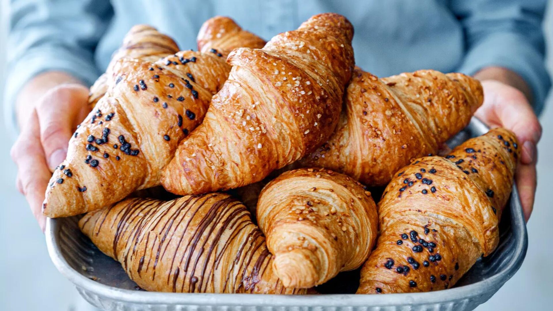 Eine Person hält ein Tablett mit verschiedenen Croissants in der Hand, von denen einige mit Schokolade, Zucker oder Körnern belegt sind und goldene, flockige Teigschichten aufweisen.