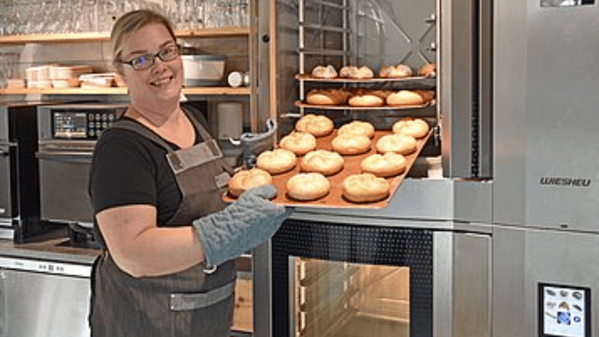 Eine lächelnde Frau mit Brille, Schürze und Ofenhandschuh hält ein Tablett mit frisch gebackenen Brötchen vor einem Industrieofen in einer modernen Küche.