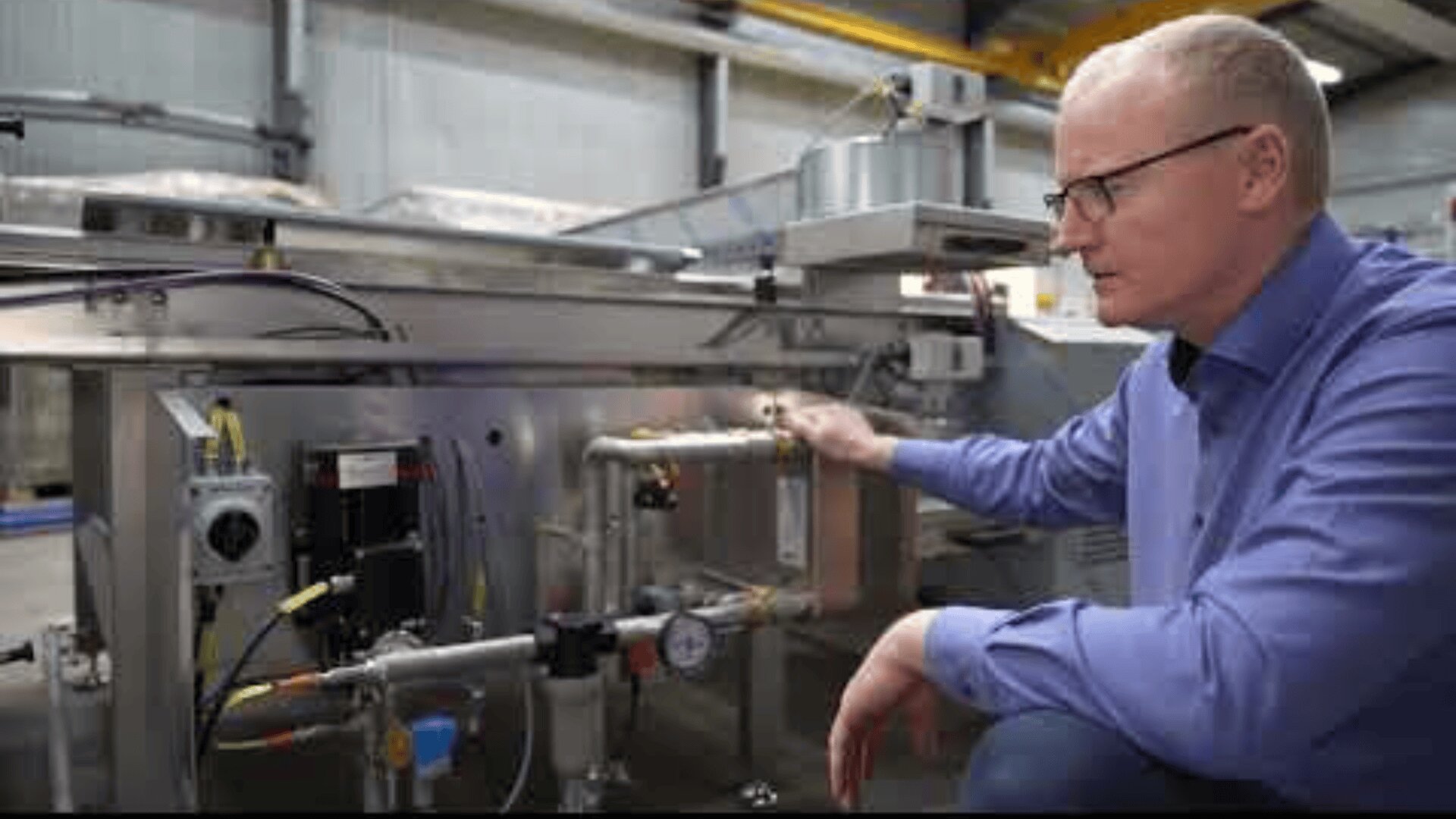 A man wearing glasses and a blue shirt examines industrial machinery in a factory setting, pointing to pipes and gauges on the equipment.