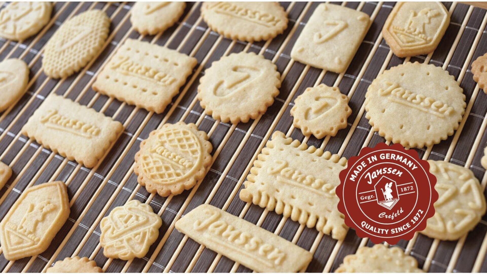 Assorted embossed butter cookies cooling on a wire rack, with a red circular “Made in Germany Janssen” quality seal logo in the lower right corner.
