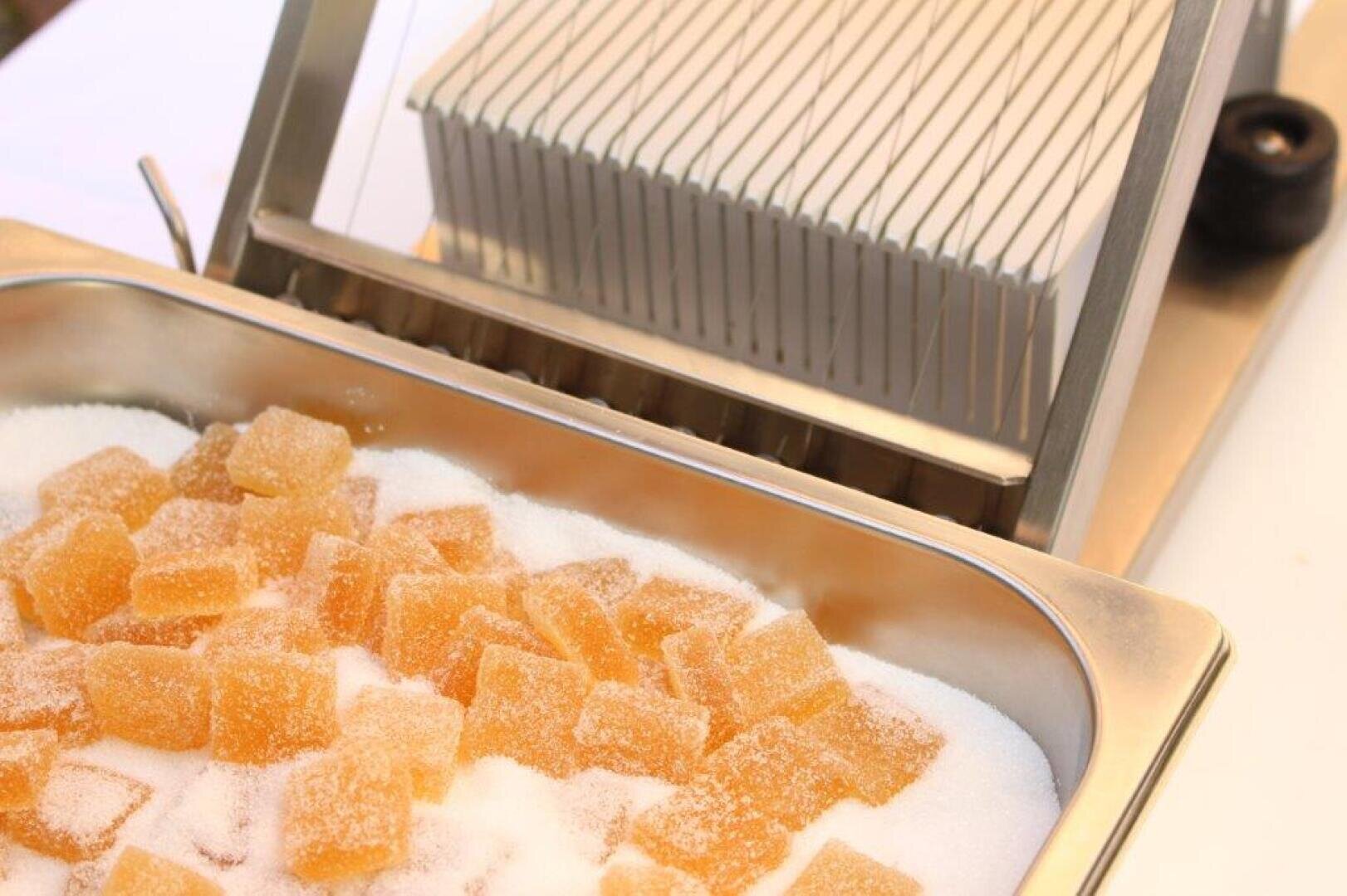 Ein Metalltablett, gefüllt mit zuckerüberzogenen gelben Geleewürfeln, steht vor einer Schneidemaschine mit gleichmäßig verteilten Drähten.