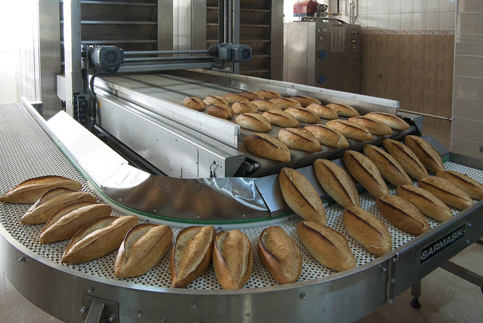 Frisch gebackene Brote laufen in einer industriellen Bäckerei über ein Förderband, im Hintergrund sind ein Ofen und gekachelte Wände zu sehen.