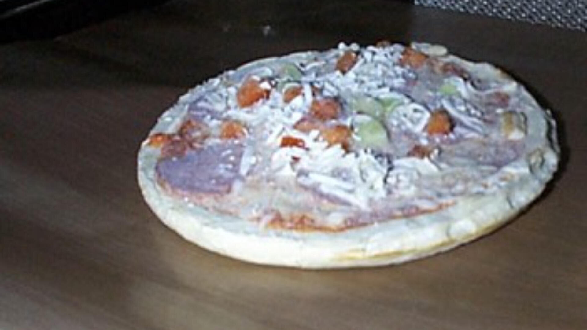 Eine gefrorene, ungebackene Pizza mit sichtbarem Käse, grünen Paprikastreifen und Fleischstücken liegt auf einem braunen Ofenrost.