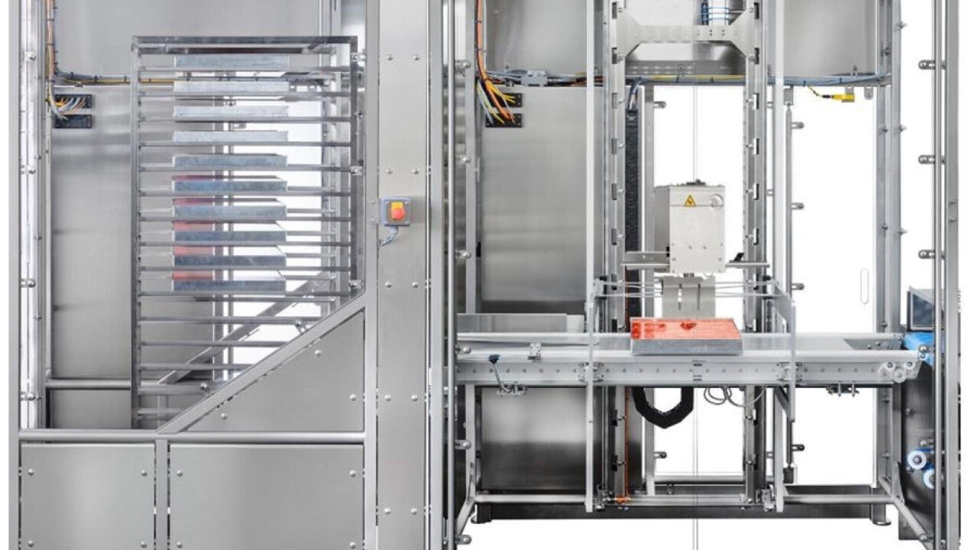 Industrial packaging machine made of stainless steel, with conveyor belts, wiring, and control panels visible. An orange item is being processed in the center. The setup appears clean and organized, suitable for automated production.