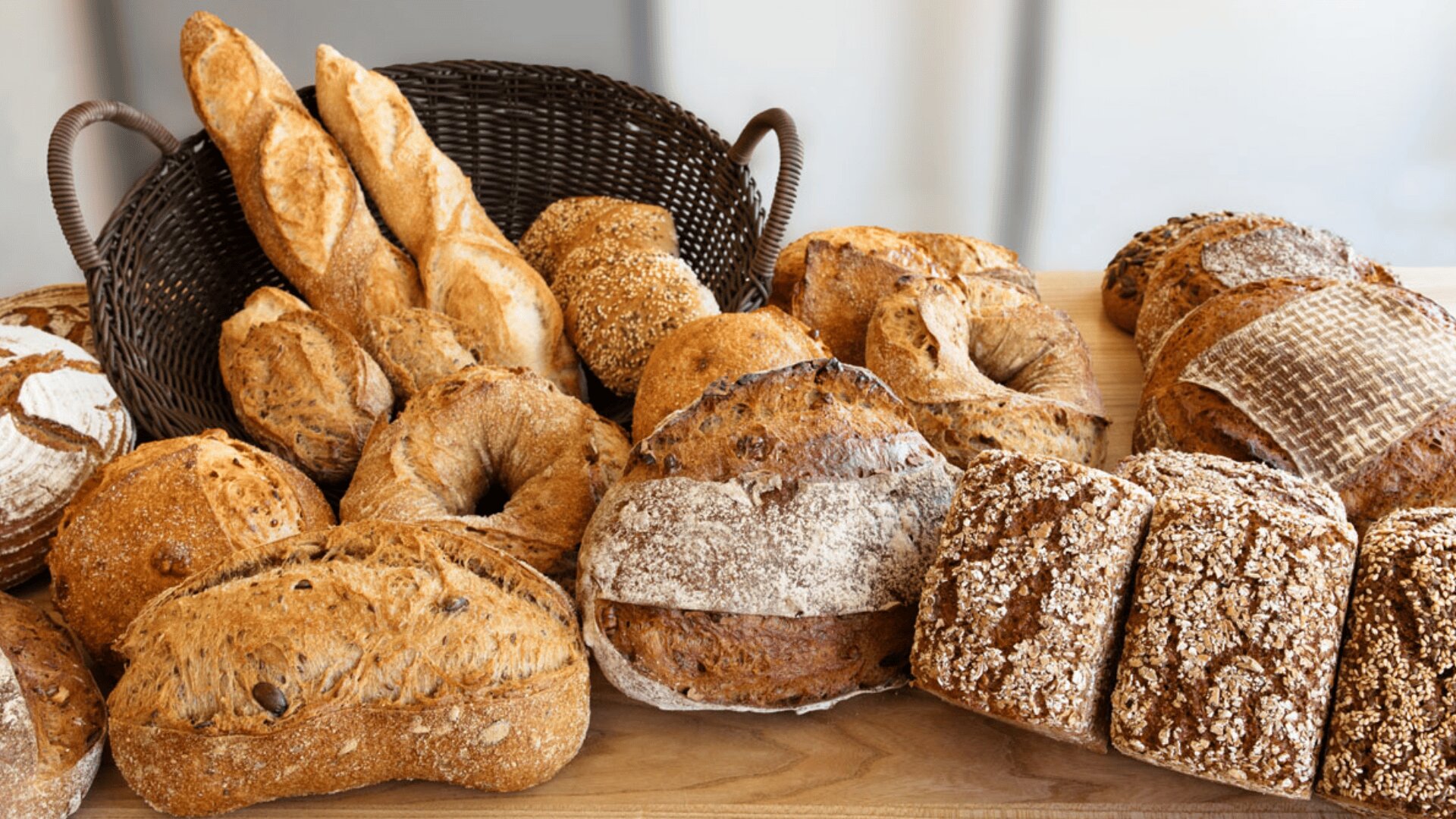 Auf einer Holzoberfläche mit einem Weidenkorb im Hintergrund werden verschiedene handwerklich hergestellte Brote präsentiert, darunter Baguettes, runde Brote, Brötchen und Brote mit Körnern.