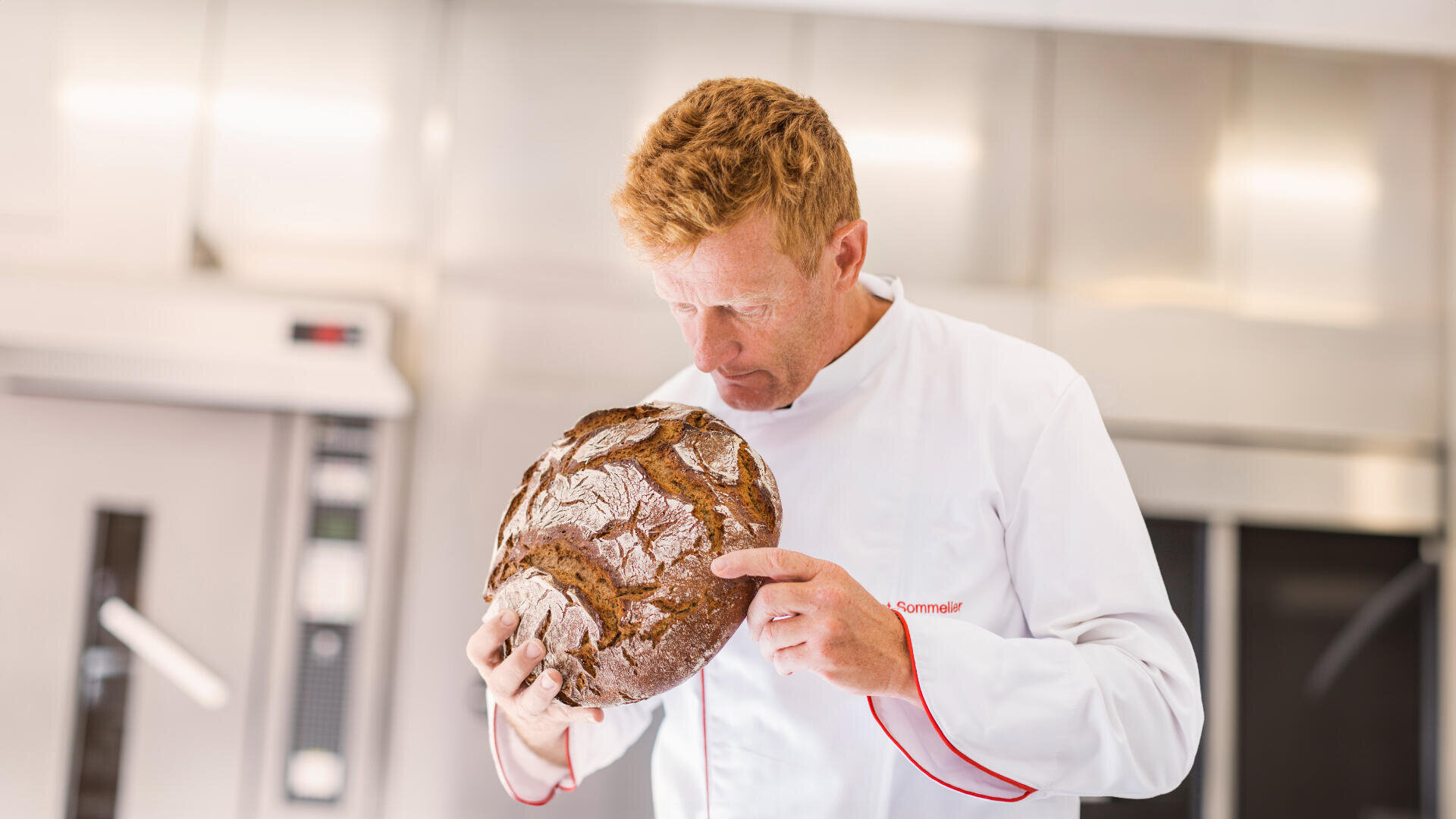 Ein Bäcker mit kurzen roten Haaren und einem weißen Kittel mit rotem Besatz steht in einer modernen Küche und betrachtet einen runden, rustikalen Laib Brot.