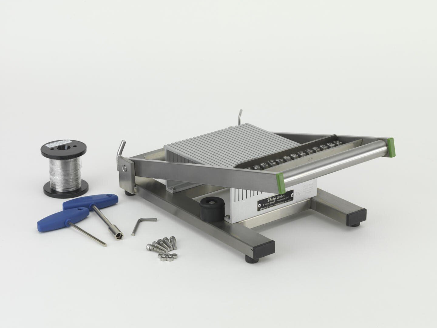 Auf einer weißen Fläche steht eine Metalldrahtschneidemaschine, daneben sind eine Drahtspule, ein Werkzeug mit blauem Griff, Schrauben und kleine Metallteile angeordnet.