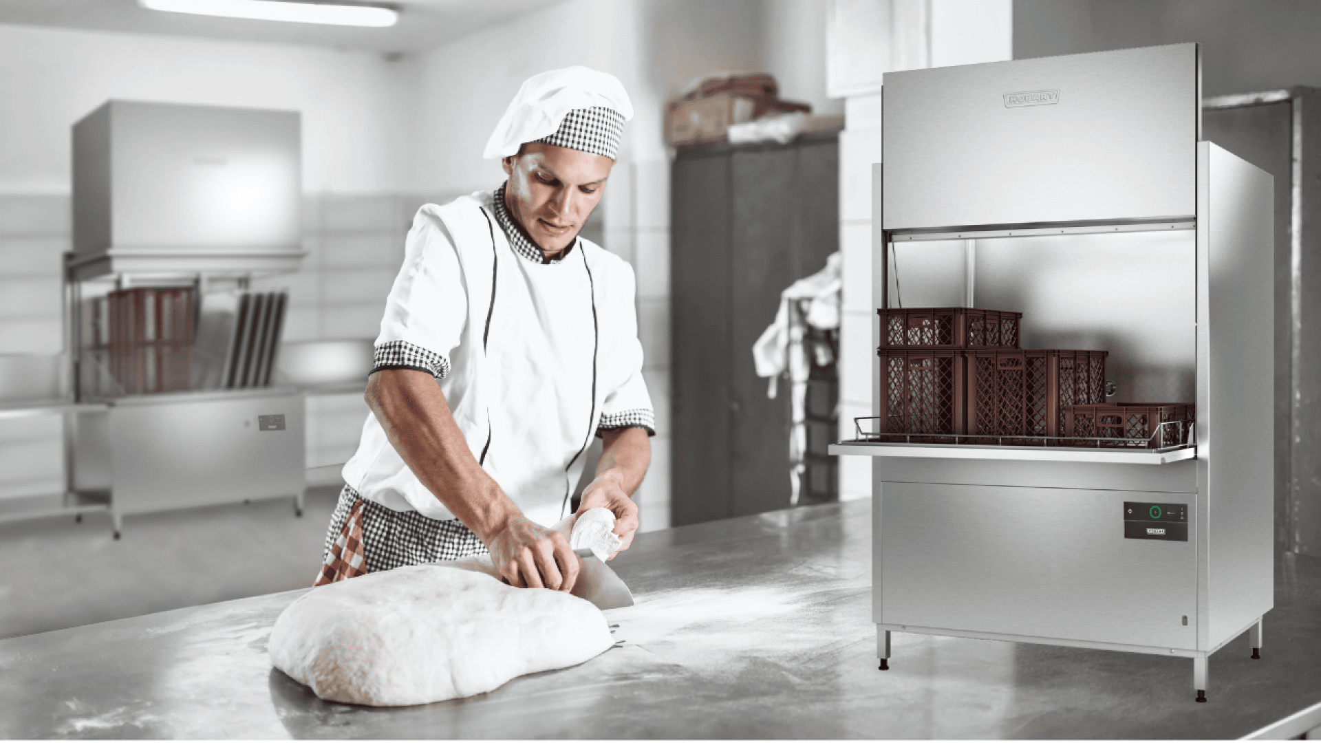Ein Bäcker in weißer Uniform und mit Hut knetet den Teig auf einer bemehlten Arbeitsfläche in einer Bäckerei. Auf der rechten Seite des Bildes ist eine große Spülmaschine aus Edelstahl mit braunen Kisten zu sehen.