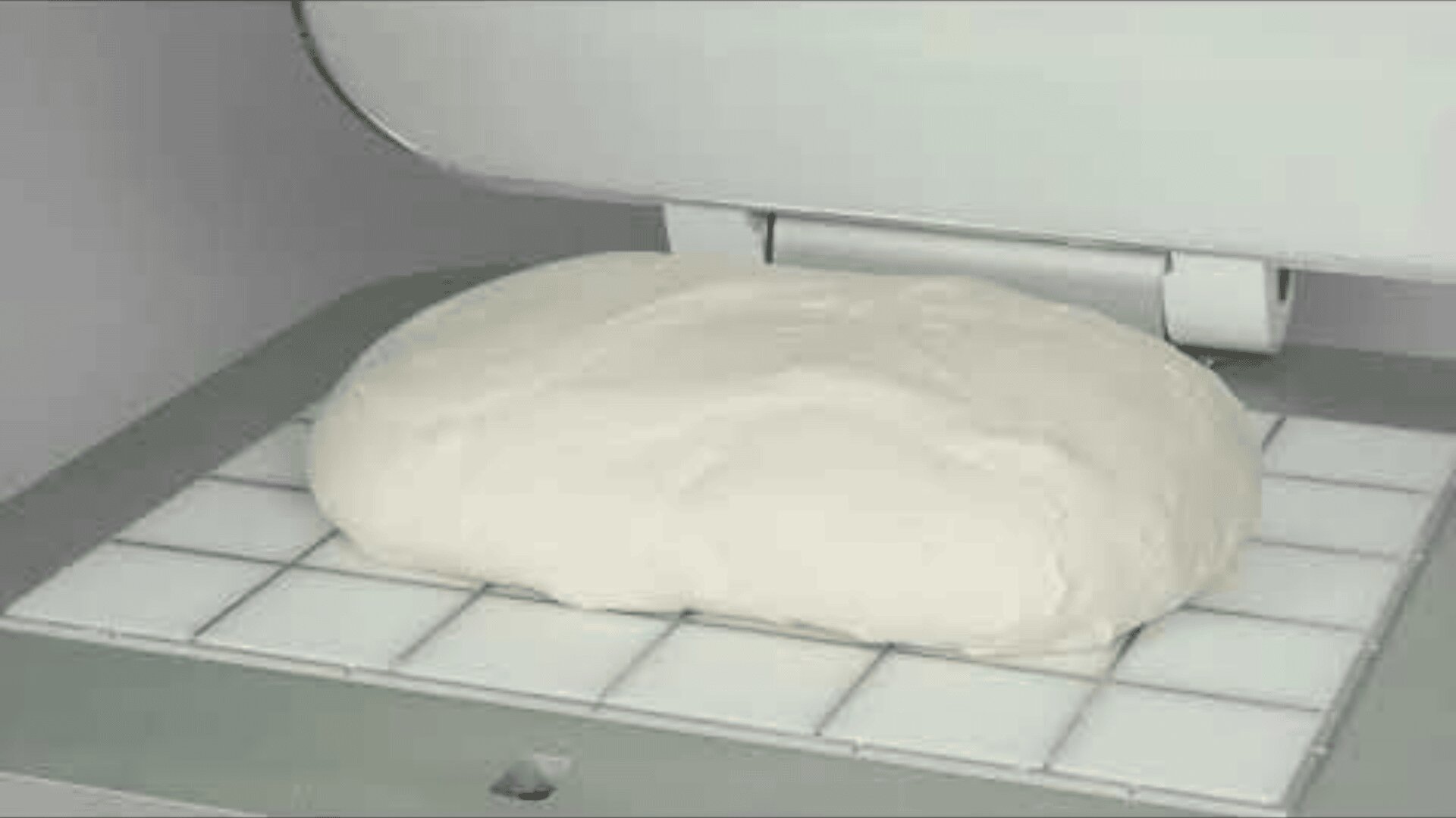 Ein großes Stück Brotteig ruht auf einer gitterartigen Oberfläche unter einem teilweise geöffneten Deckel einer Industriemaschine, möglicherweise zum Kneten oder Pressen.