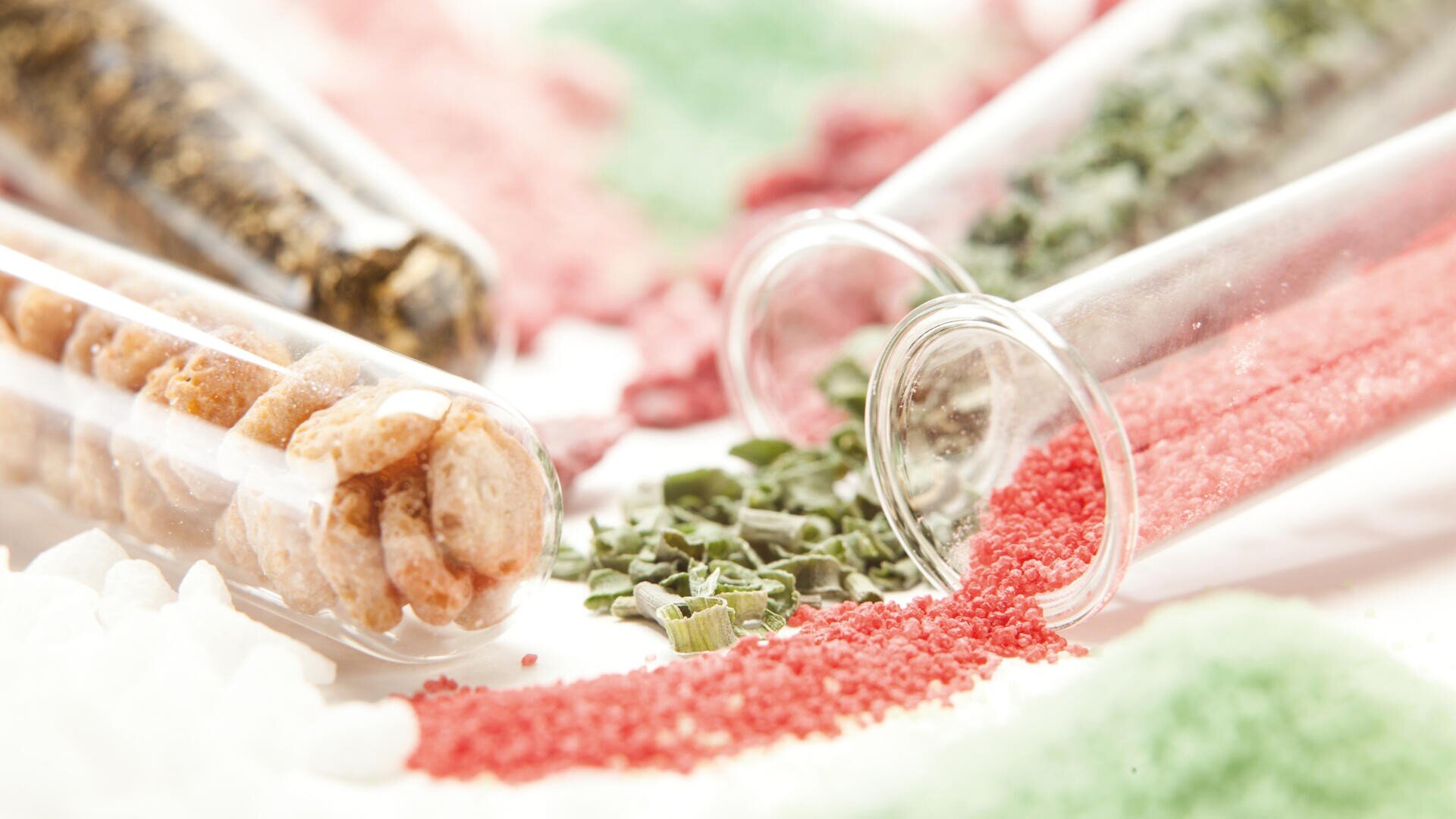 Auf einer weißen Fläche liegen mehrere Glasreagenzgläser, aus denen bunte Inhalte wie rotes, grünes und weißes Granulat sowie getrocknete Kräuter und runde Kügelchen austreten. Die Körnchen und Kräuter bilden bunte, verstreute Haufen.