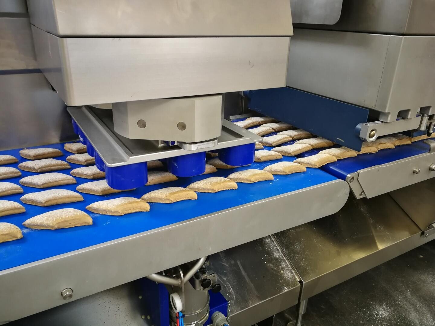 Eine Lebensmittelproduktionsmaschine formt Teigstücke auf einem blauen Förderband in einer Fabrikhalle und bereitet sie zum Backen oder zur Weiterverarbeitung vor.