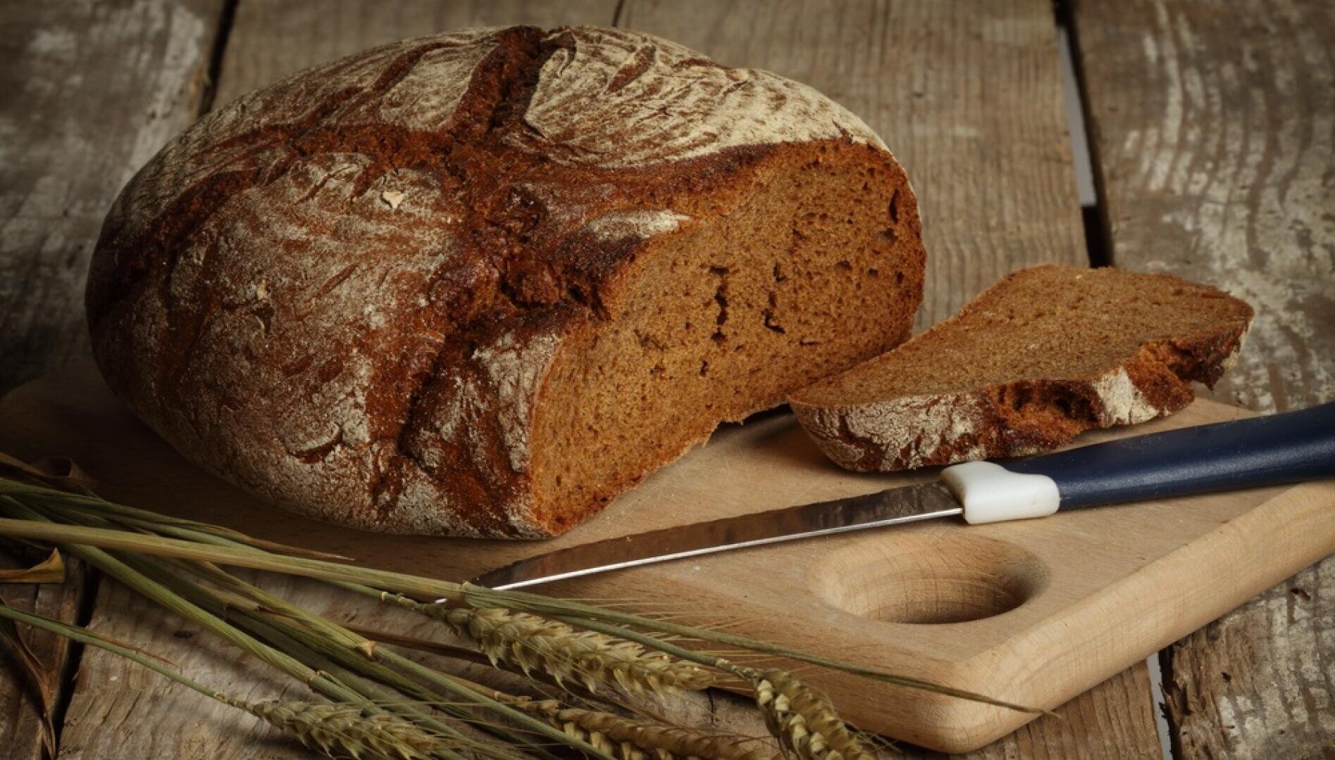 Ein runder Laib rustikales Schwarzbrot mit einer abgeschnittenen Scheibe liegt neben einem Brotmesser auf einem hölzernen Schneidebrett. Auf dem Brett liegen auch Weizenhalme, und der Hintergrund besteht aus verwittertem Holz.