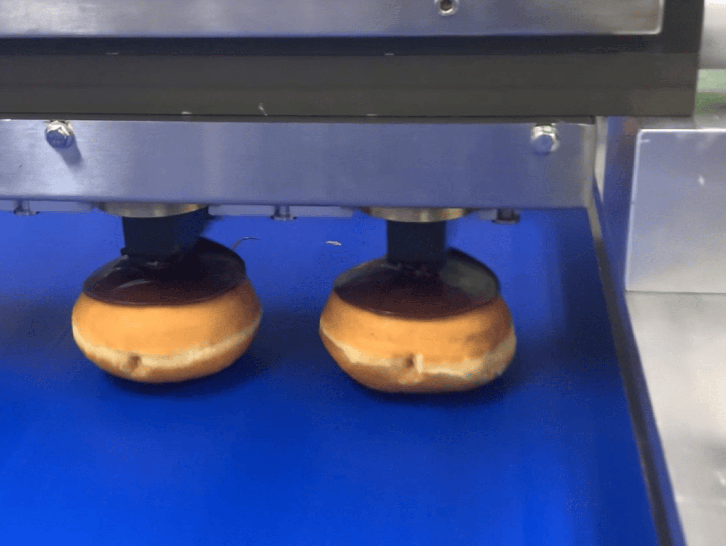 Two doughnuts on a blue conveyor belt are being topped with chocolate glaze by a machine, as part of an automated production process.