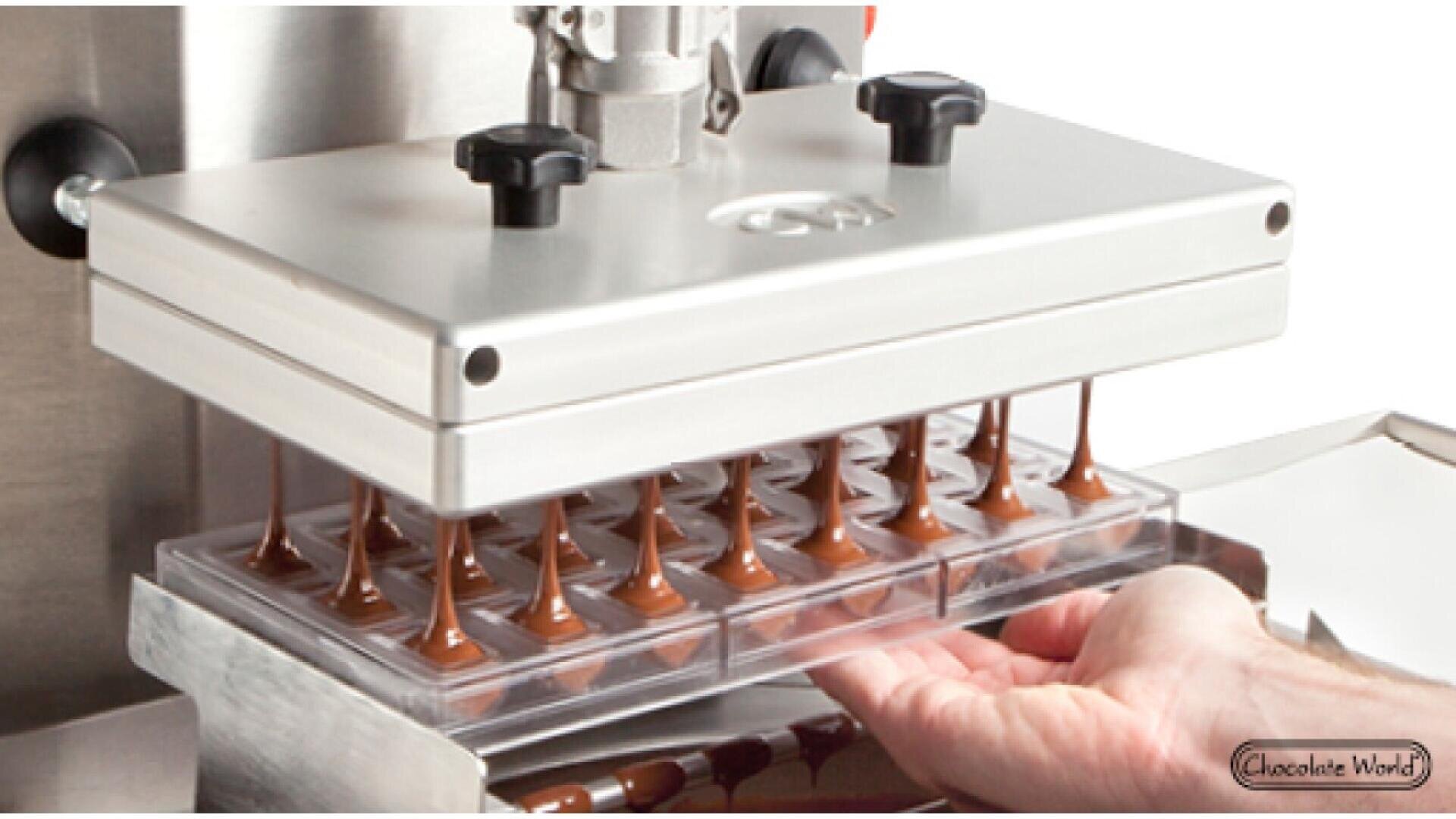 A chocolate tempering machine fills a mold with liquid chocolate, creating rows of chocolates, while a persons hand steadies the mold underneath the dispenser.