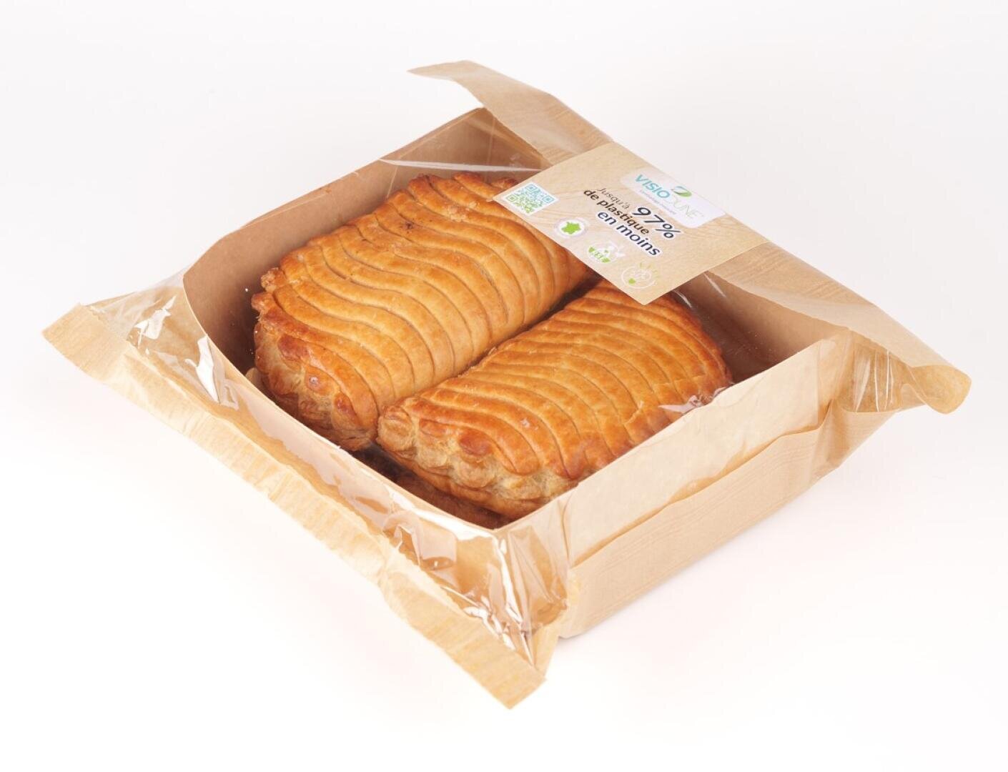 Two golden-brown puff pastry turnovers are displayed in a partially open, brown paper and clear plastic bakery box on a white background. A label with green and blue accents is on top of the box.
