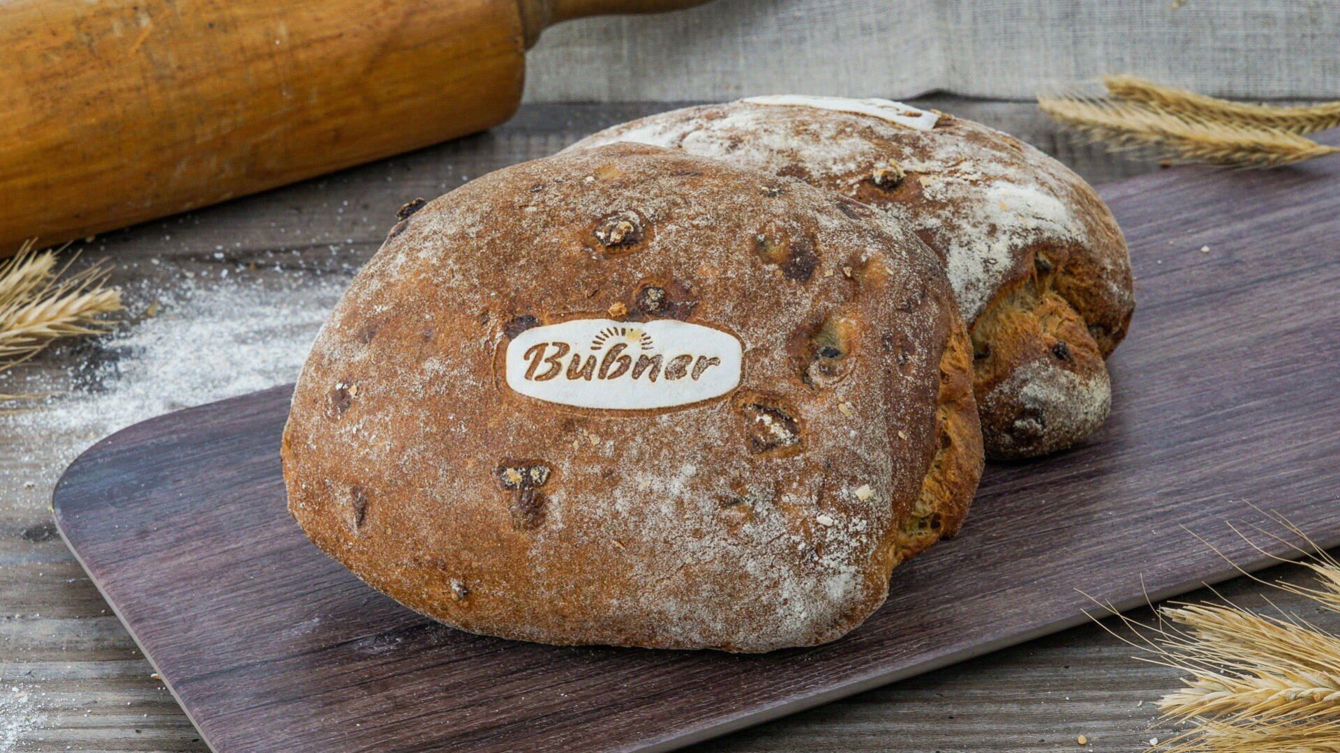 Zwei rustikale Brotlaibe mit dem Aufdruck Bubner liegen auf einem Holzbrett, umgeben von Weizenhalmen und einem Nudelholz auf einer Holzfläche.