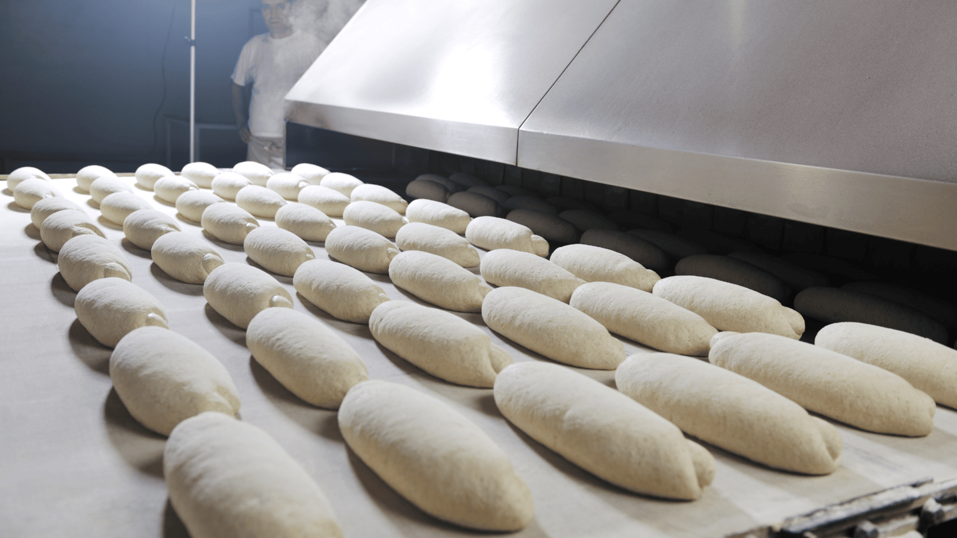 Reihen von ungebackenen Baguette-Broten sind auf einem Förderband aufgereiht und werden in einen großen Industrieofen geschoben. Eine Person in weißer Uniform steht in der Nähe im Hintergrund, teilweise durch Dampf verdeckt.