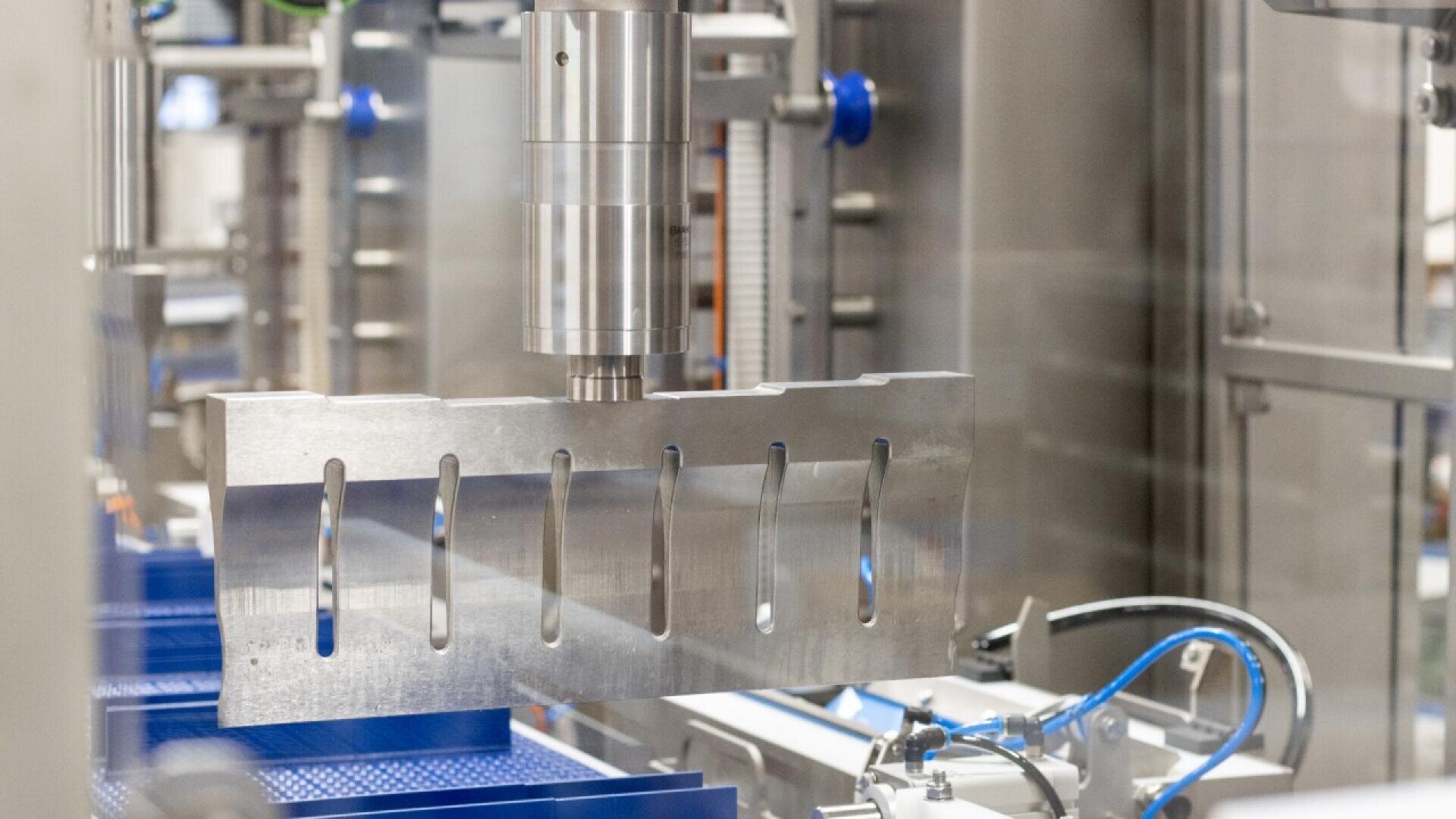 Nahaufnahme einer Industriemaschine mit einer großen Metallklinge mit mehreren rechteckigen Schlitzen, umgeben von verschiedenen Metallteilen und blauen Kabeln.