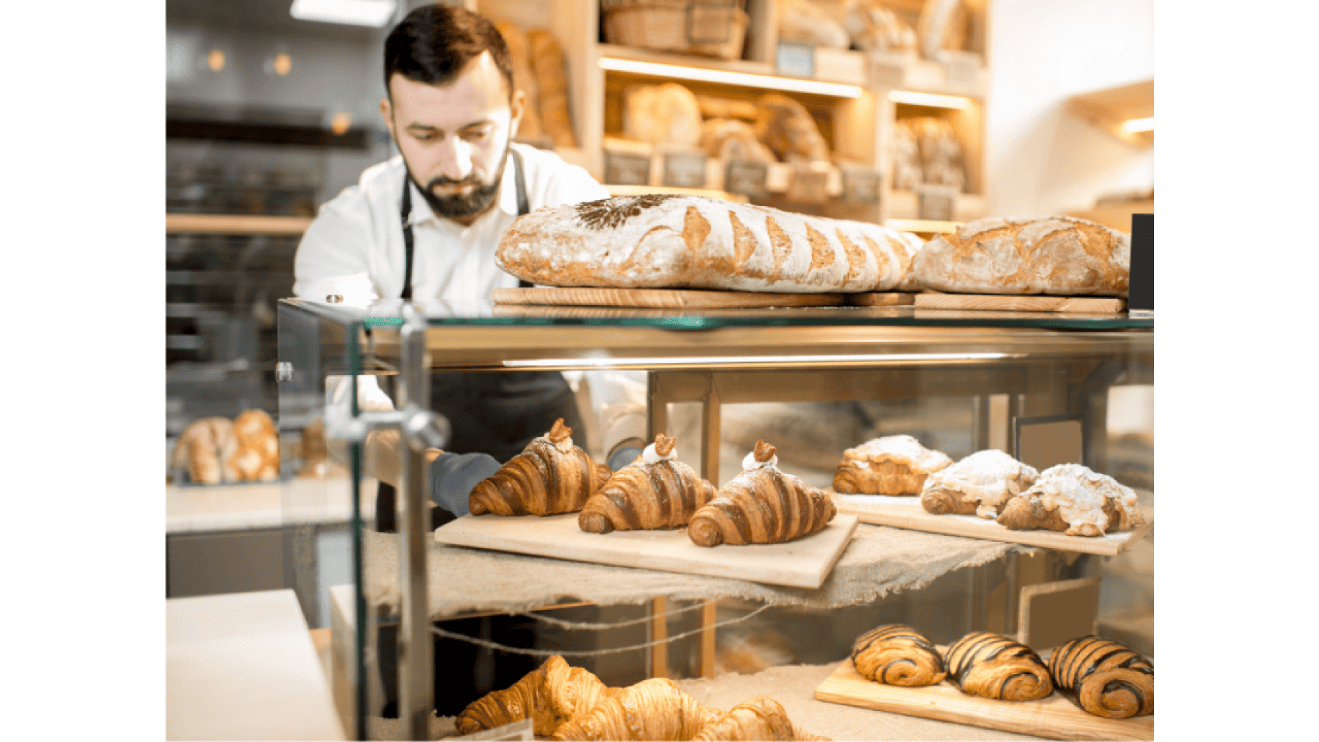 Ein Bäcker in weißem Hemd und schwarzer Schürze ordnet frische Brotlaibe hinter einer mit Croissants und Gebäck gefüllten Glasauslage in einer Bäckerei mit Holzregalen mit Brot im Hintergrund an.
