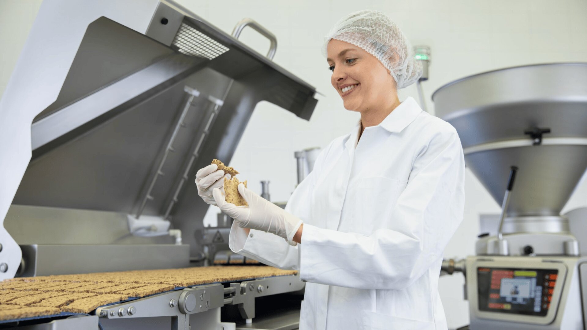 Eine Frau in weißem Laborkittel, Haarnetz und Handschuhen inspiziert einen Müsliriegel auf einer Lebensmittelproduktionslinie in einer modernen Fabrik mit Geräten aus Edelstahl.