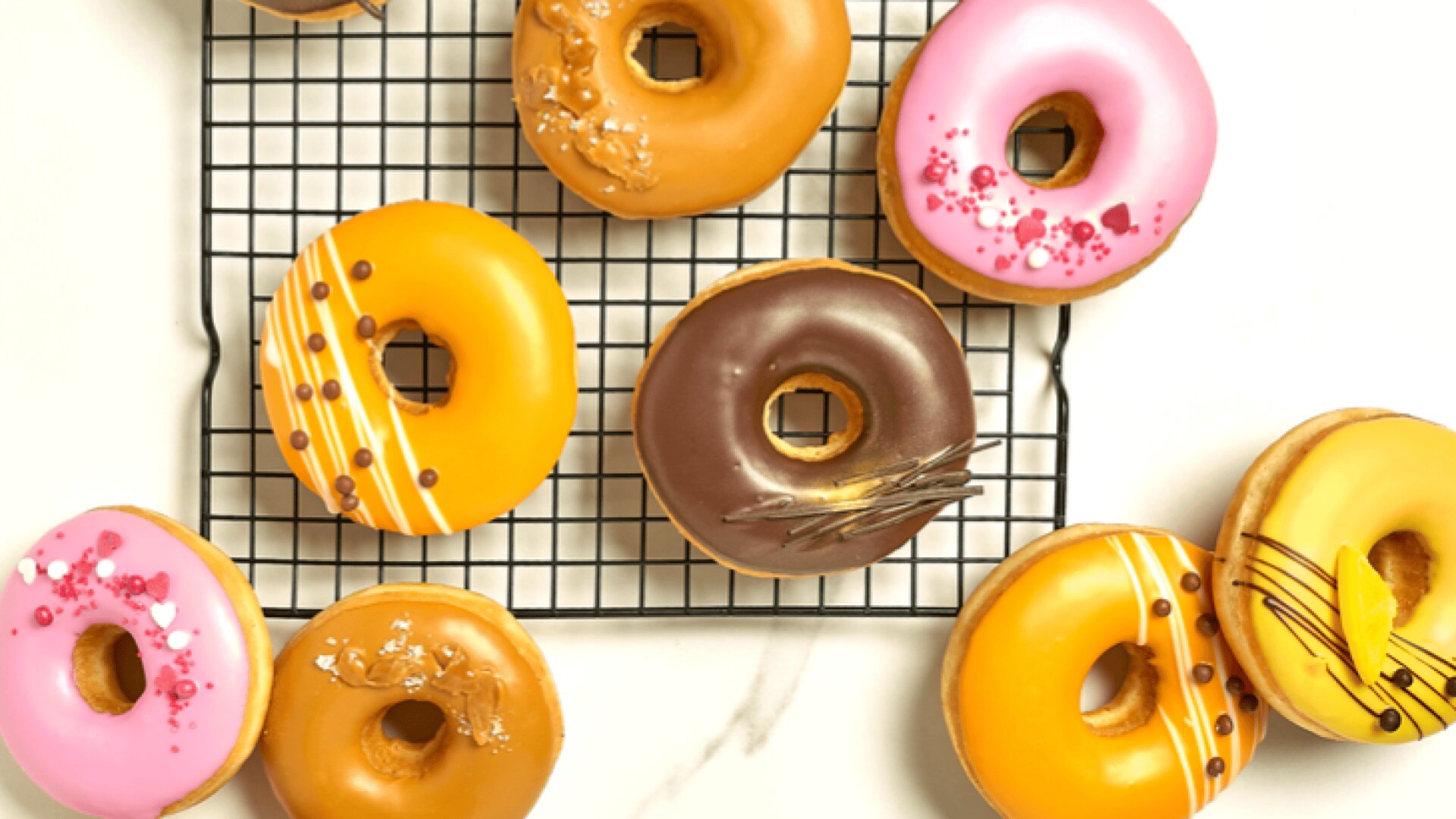 A variety of colorful, frosted donuts with decorative toppings are arranged on and around a cooling rack atop a white marble surface.