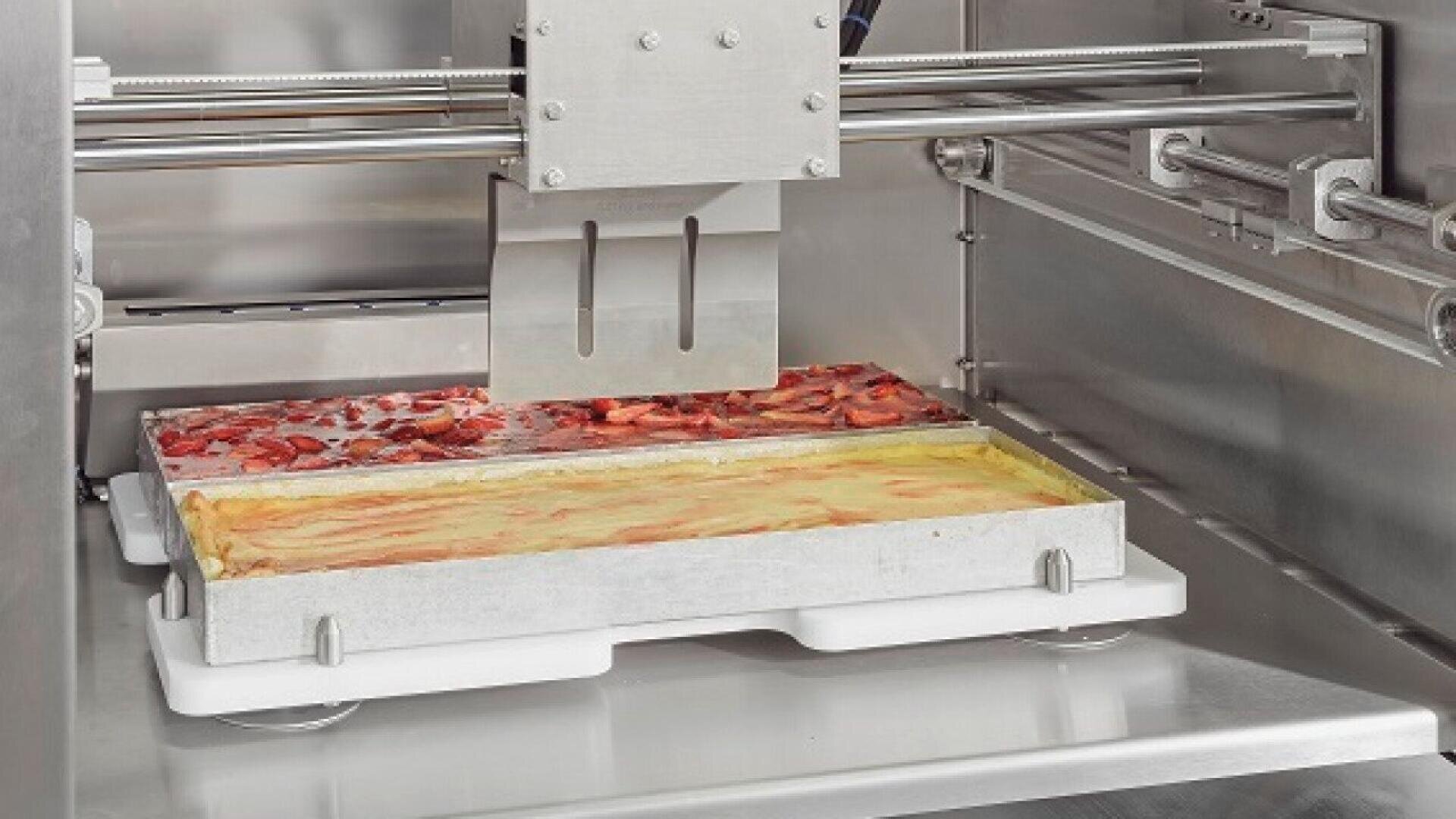 A close-up of a 3D printer preparing two rectangular layered desserts, one topped with strawberries and the other with a yellow custard, inside a stainless steel machine.