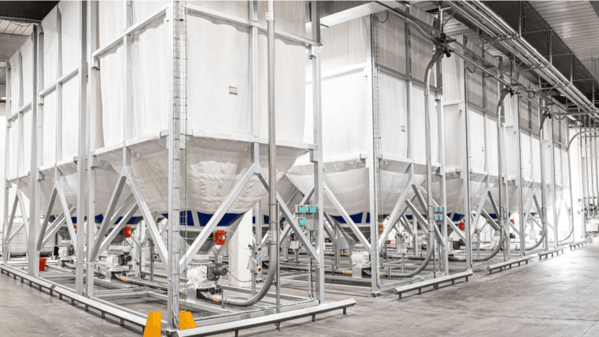 Große industrielle Lagersilos mit Metallrahmen und weißen Stoffwänden sind in einem geräumigen, gut beleuchteten Lager aufgereiht. Rohre und Maschinen sind an jede Einheit angeschlossen, und der Boden ist sauber und aufgeräumt.