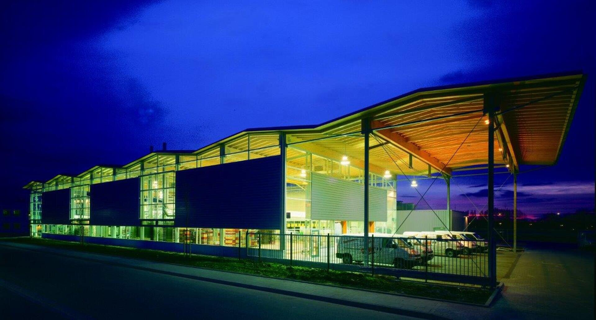 A modern building with large glass windows and an angular, overhanging roof is brightly lit at dusk. Several vehicles are parked in front, and the interior lighting creates a striking contrast with the dark blue sky.