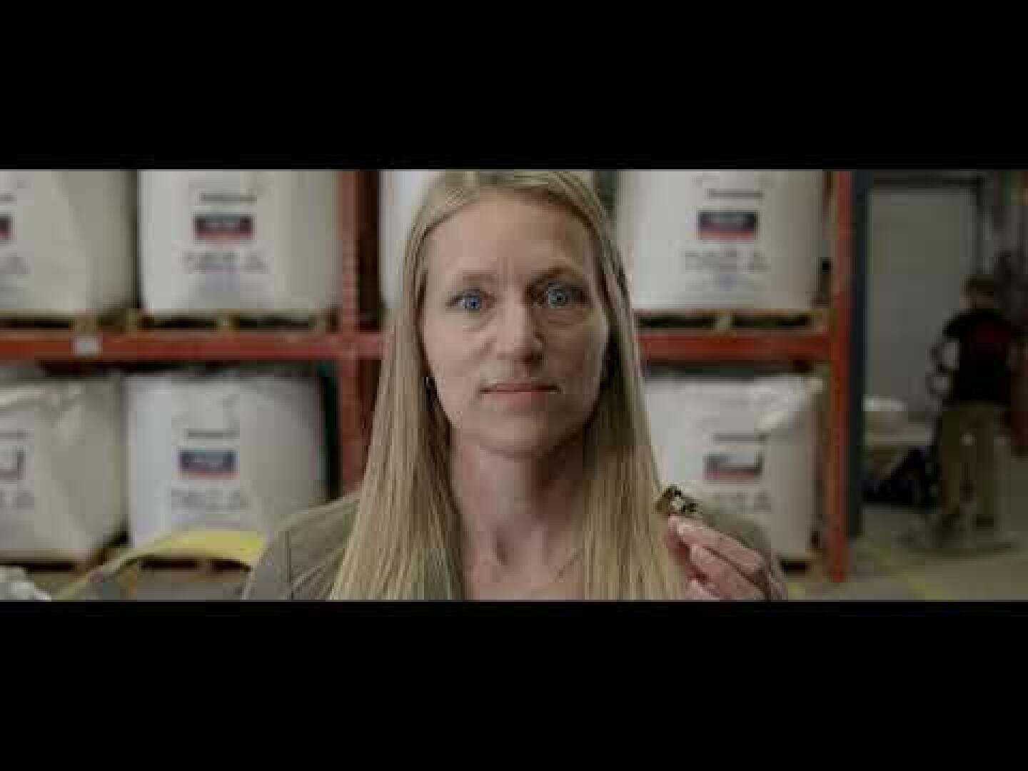 A woman with long blonde hair stands indoors holding a small object, possibly a nut or bolt, in her hand. Large industrial bags are stacked on shelves behind her in a warehouse setting.