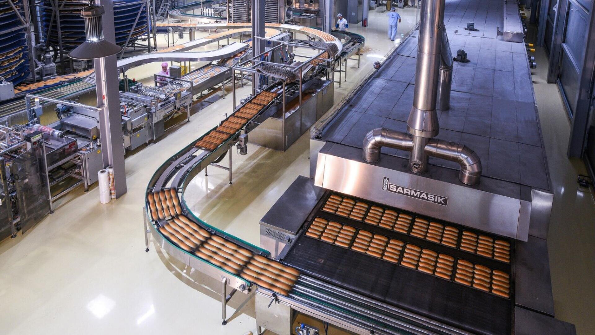 Reihen von Brotlaiben bewegen sich auf einem Förderband in einer großen industriellen Bäckerei durch Maschinen und Öfen aus rostfreiem Stahl. Im Hintergrund sind Arbeiter in Schutzkleidung zu sehen.