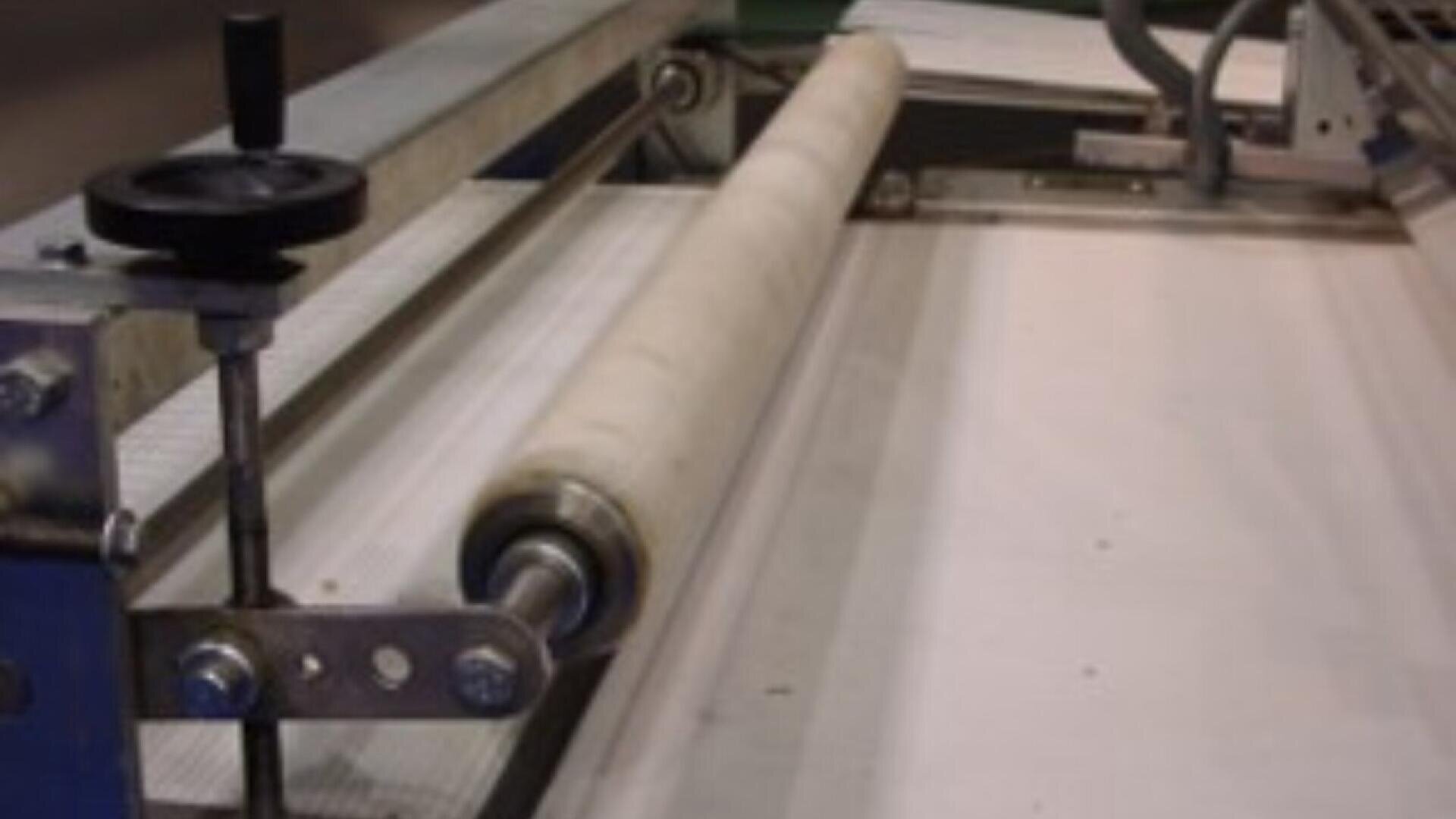A close-up view of a fabric being rolled through an industrial machine with metal rollers and a crank handle, likely part of a textile manufacturing or processing operation.