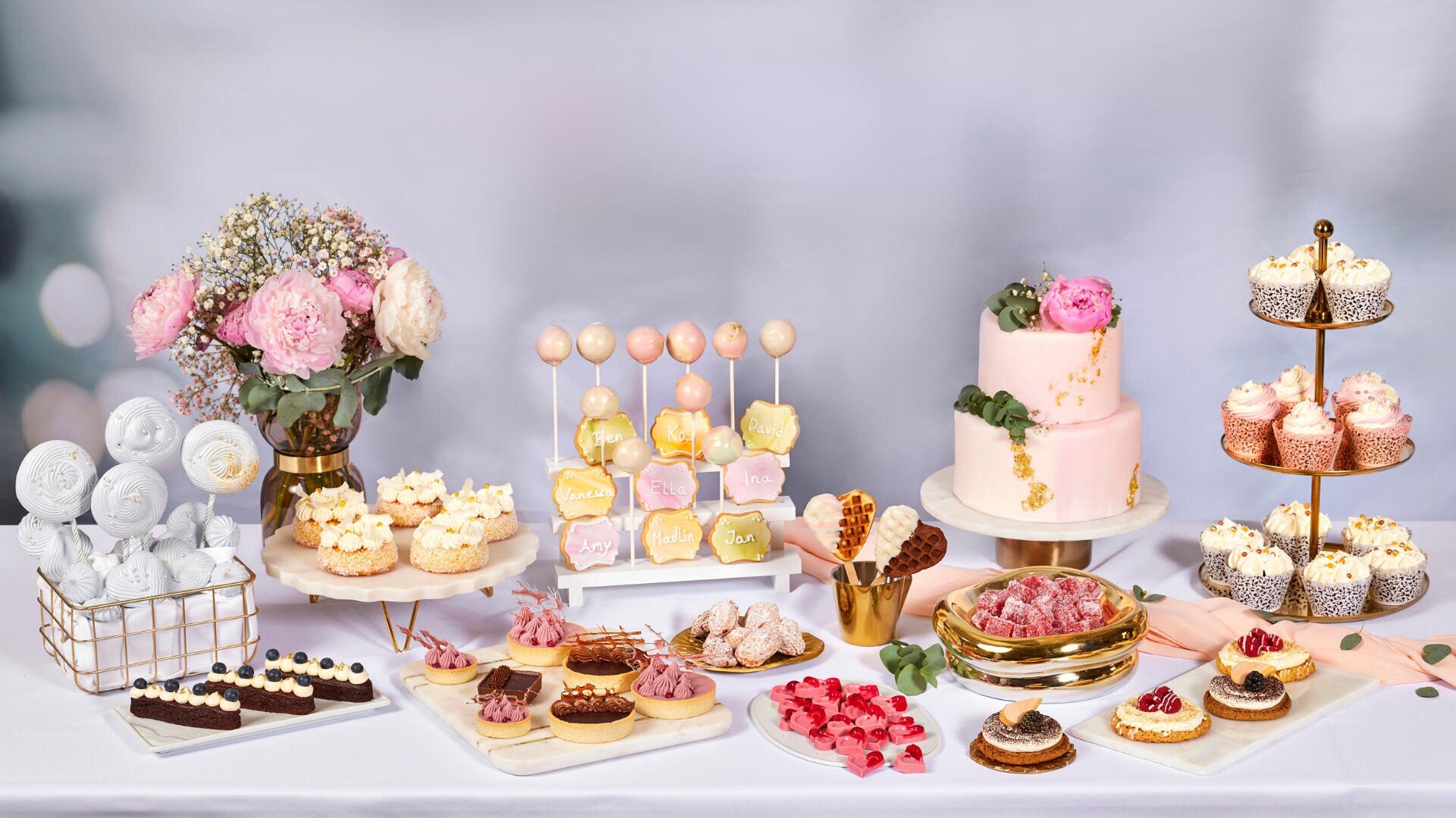 Ein mit Blumen dekorierter Desserttisch mit einer rosa Blumentorte, Cupcakes, Cake Pops, Keksen, Brownies, Törtchen und Süßigkeiten, die auf Tabletts, Ständern und Tellern vor einem sanften Hintergrund angeordnet sind.