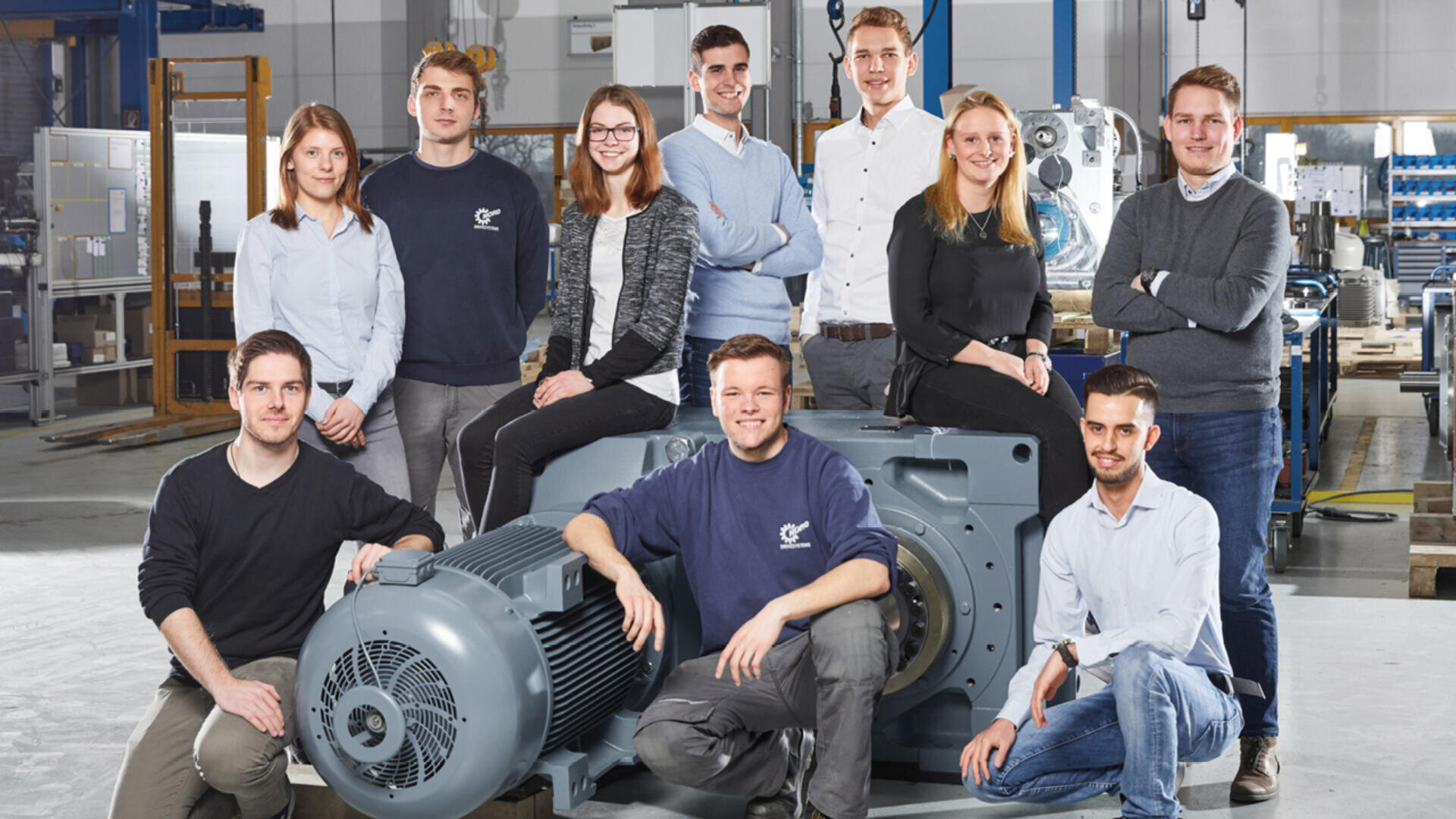Eine Gruppe von zehn leger gekleideten jungen Erwachsenen posiert und lächelt gemeinsam in einer industriellen Umgebung. Sie stehen und sitzen um eine große graue Maschine herum. Im Hintergrund sind Werkstattausrüstung und Maschinen zu sehen.
