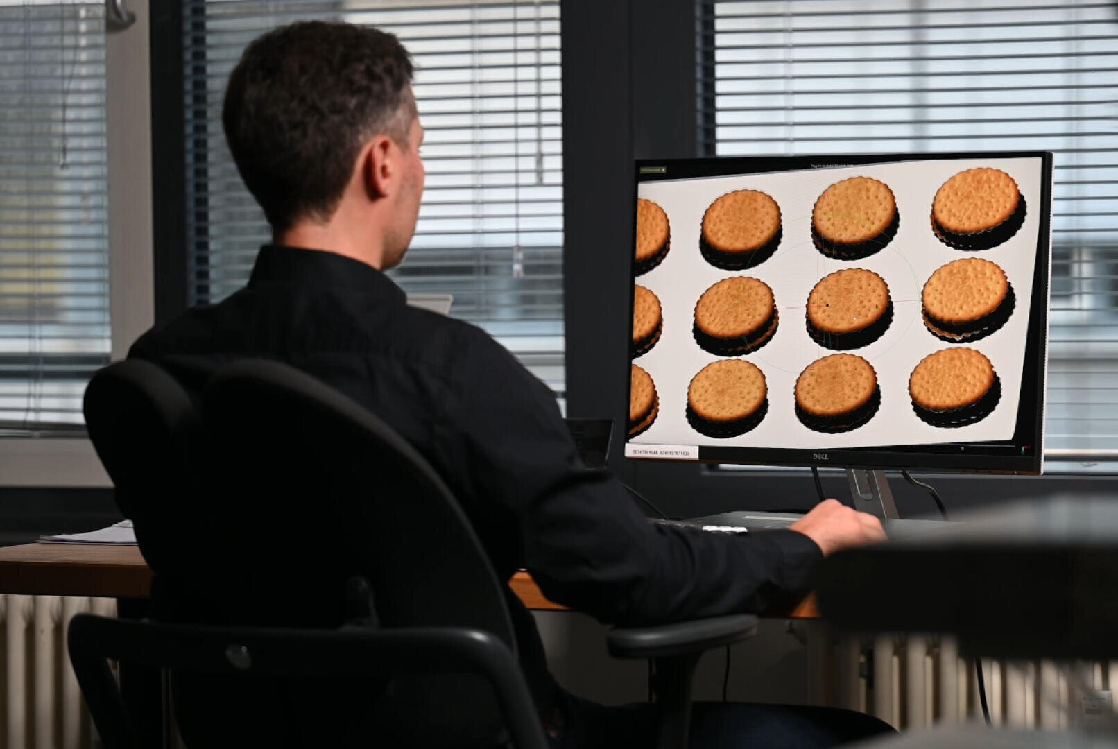 Eine Person sitzt an einem Schreibtisch mit Blick auf einen Computermonitor, auf dem Bilder von zwölf runden, goldenen Keksen in Reihen angeordnet sind. Die Fenster im Hintergrund sind mit Jalousien abgedeckt, und man kann die Büroausstattung sehen.