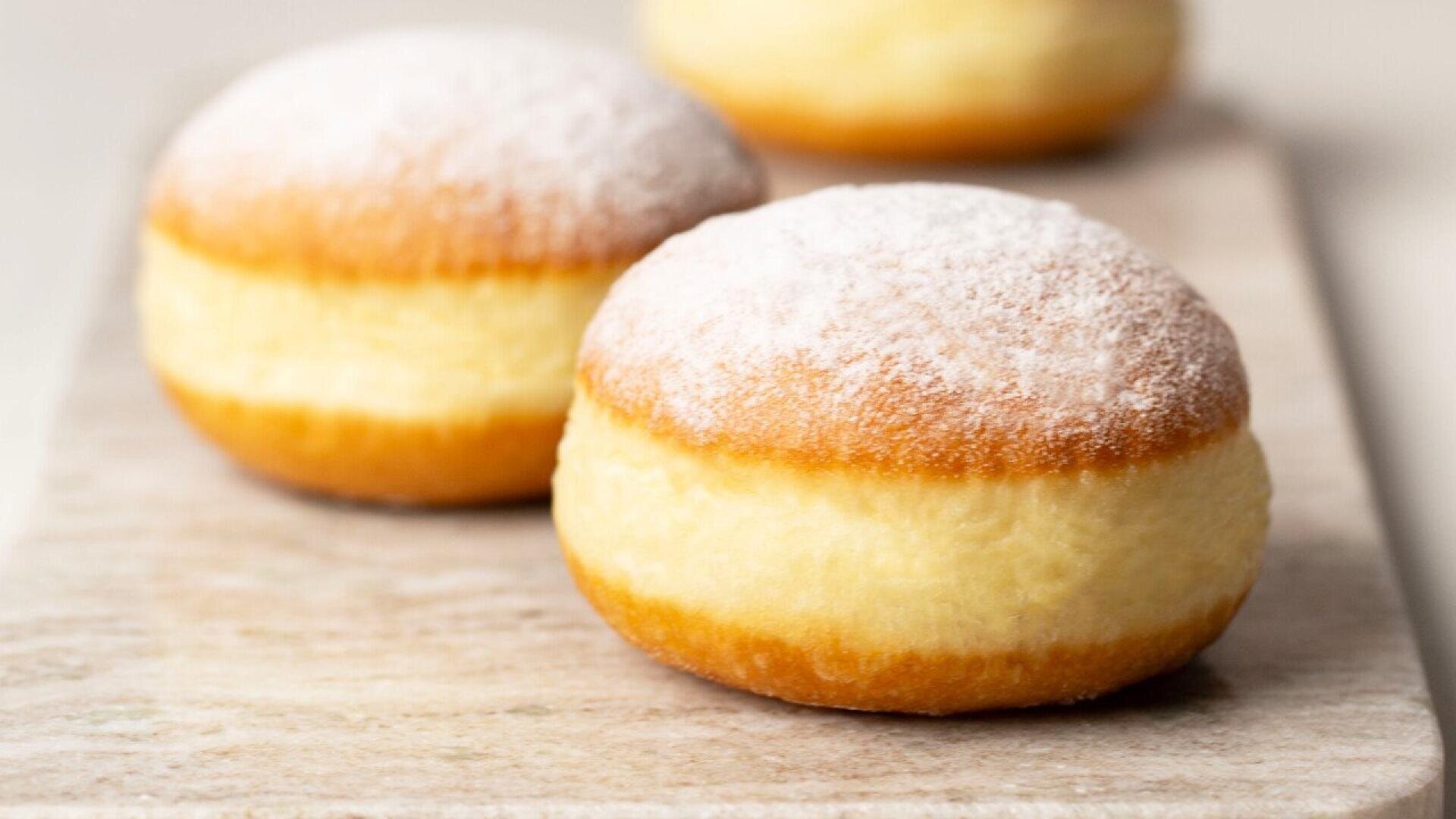 Close-up of three round, golden-brown doughnuts dusted with powdered sugar, placed on a light-colored surface. The focus is on the front doughnut, with the others slightly blurred in the background.