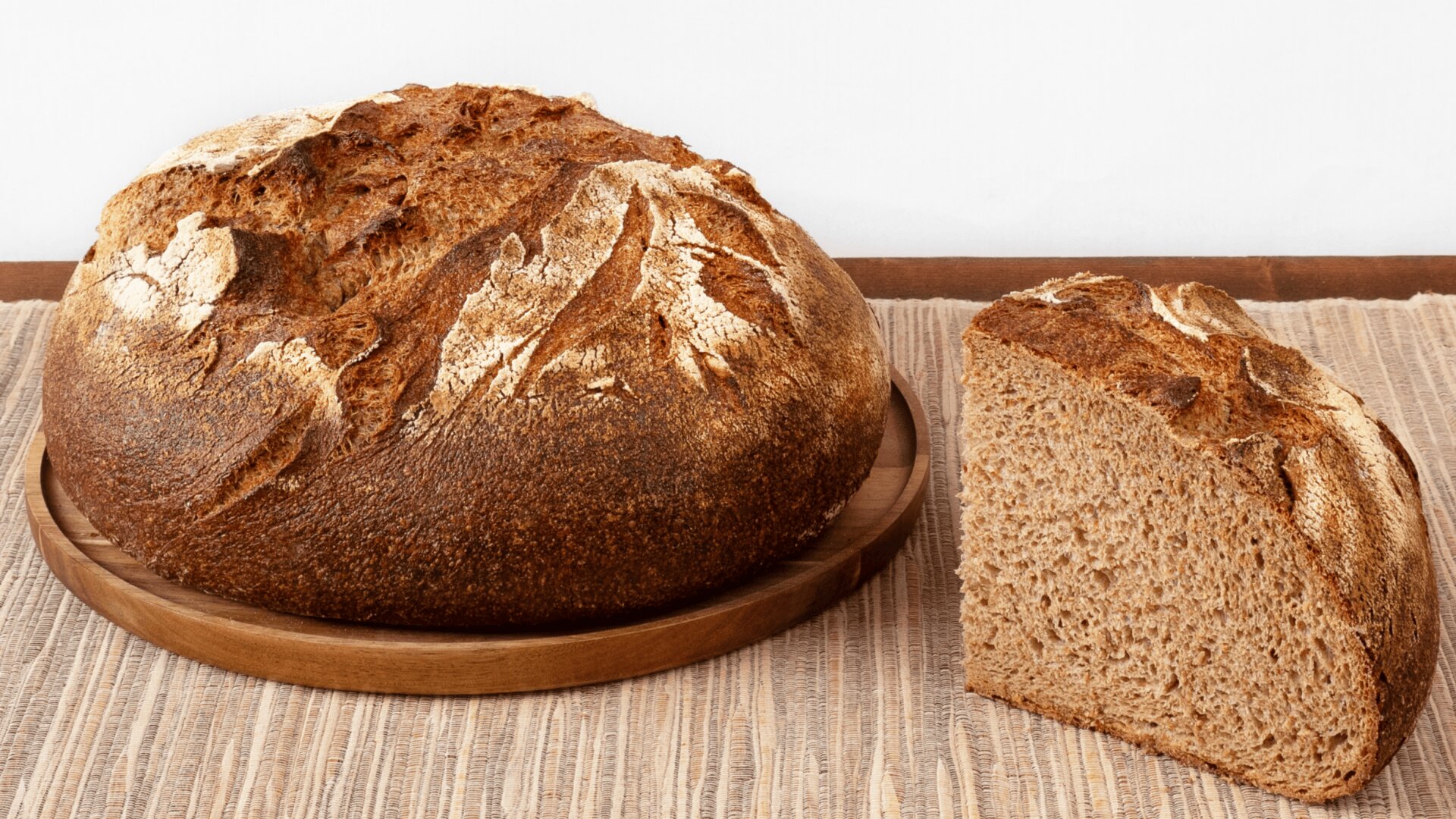 A round loaf of rustic bread sits on a wooden plate, with a single slice cut and placed beside it on a woven placemat atop a wooden table. The background is plain and white.