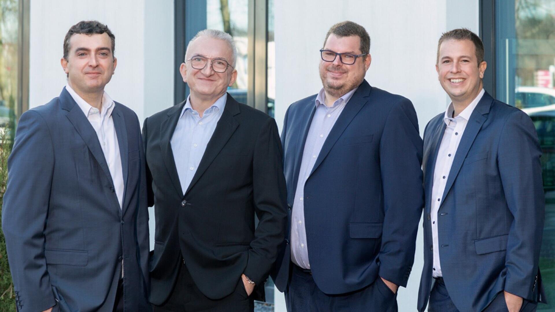 Four men wearing suits stand side by side in front of a building, smiling at the camera. They appear to be colleagues or business professionals.