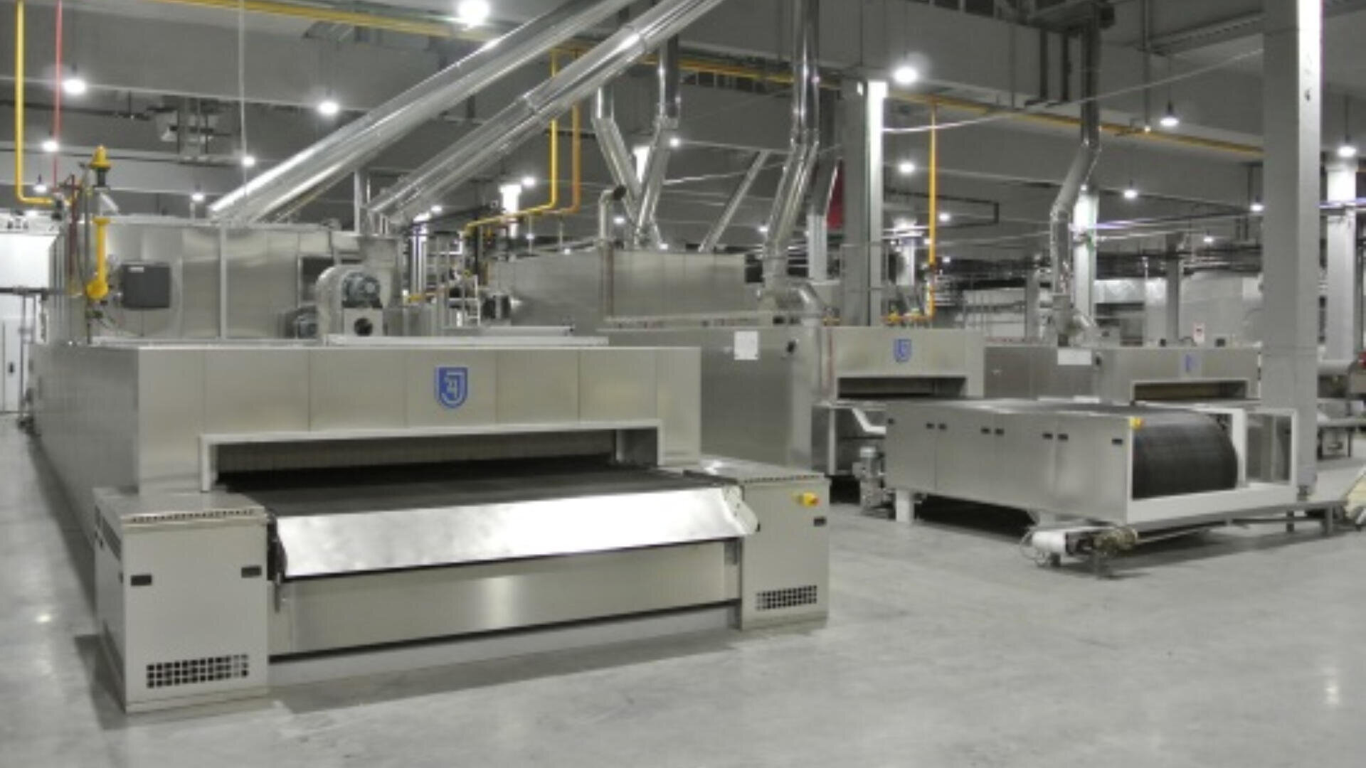 A large industrial food processing machine with conveyor belts and metal surfaces inside a spacious, well-lit factory setting. Various pipes and equipment are visible in the background.