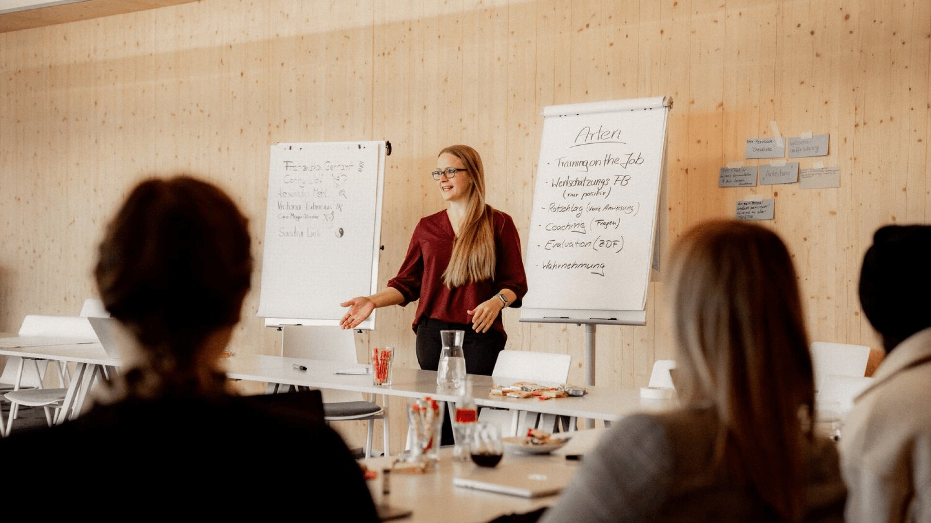 Eine Frau steht vor zwei Flipcharts und spricht zu einer Gruppe von sitzenden Personen in einem hellen Klassenzimmer. Sie gestikuliert während ihres Vortrags, und auf den Tischen stehen Notebooks, Getränke und Snacks.
