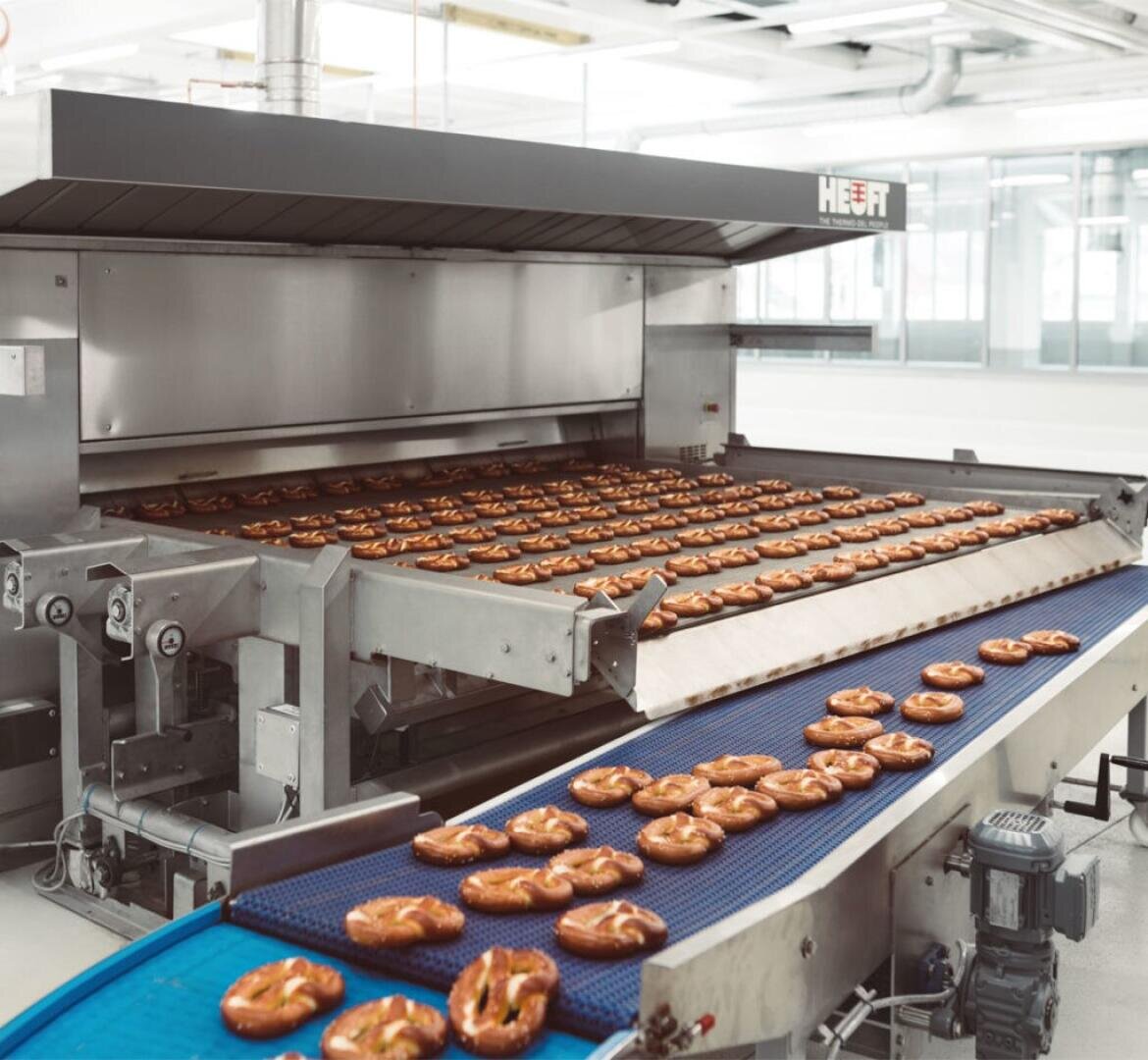 Großer Industrieofen mit mehreren Regalen mit frisch gebackenen Brezeln, die auf einem Förderband in einer hellen, modernen Bäckerei laufen. Die Brezeln sind goldbraun und gleichmäßig geformt.