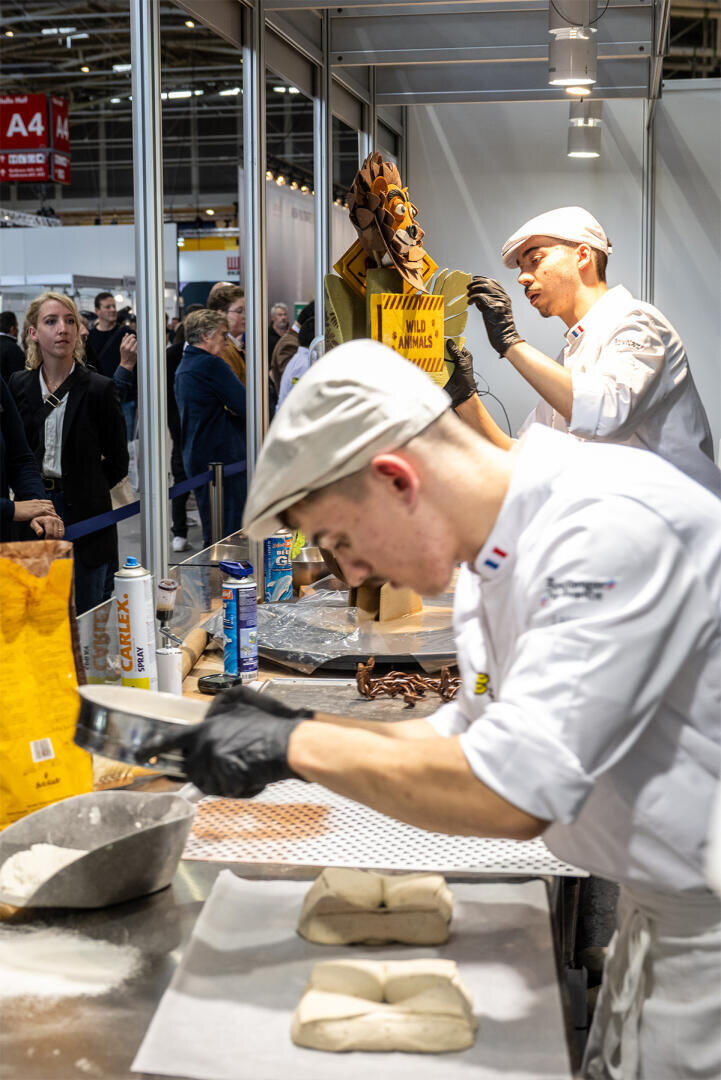 Zwei Bäcker in weißen Uniformen und Mützen arbeiten an einer Theke, formen Teig und handhaben Backutensilien, während ihnen hinter ihnen bei einer geschäftigen Veranstaltung oder Ausstellung in einem geschlossenen Raum eine Menschenmenge zusieht.