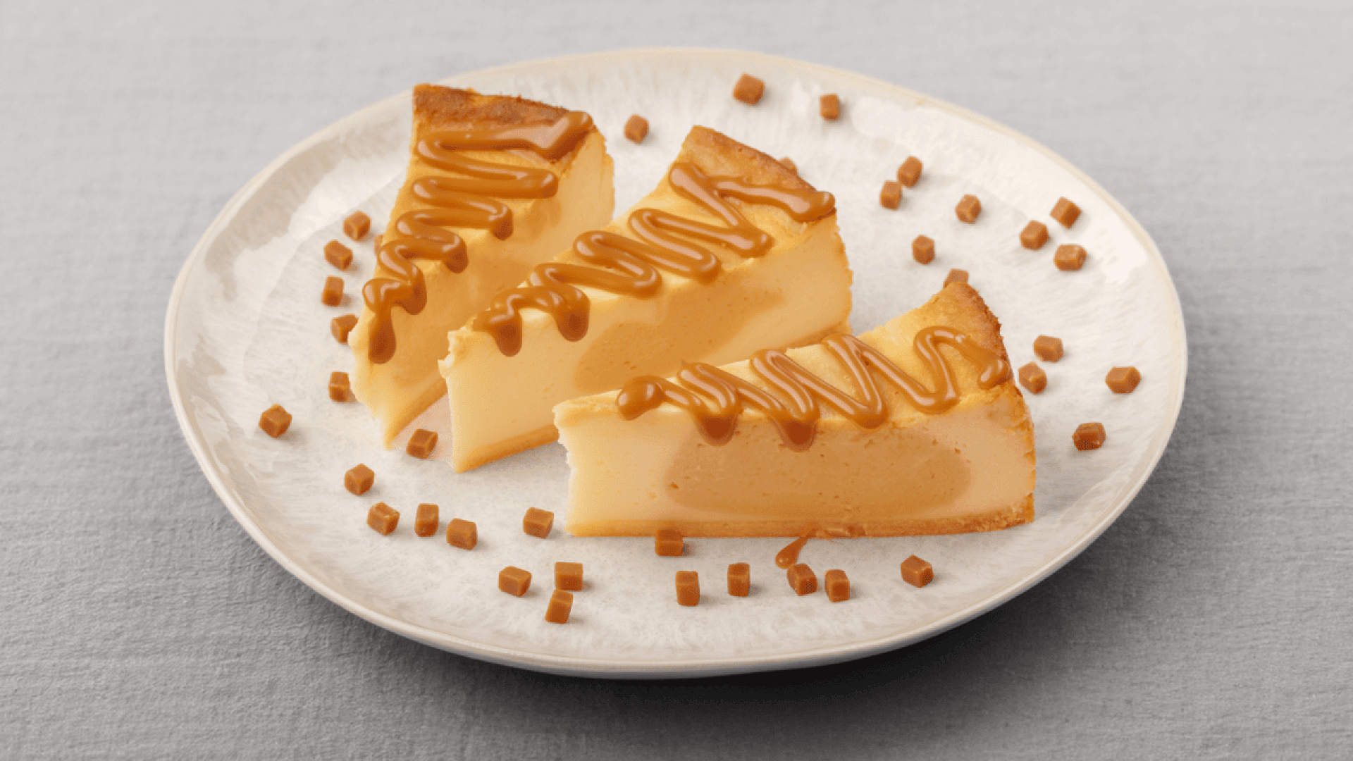 Three slices of caramel flan topped with caramel drizzle are arranged on a white plate, garnished with small caramel cubes on a light grey surface.