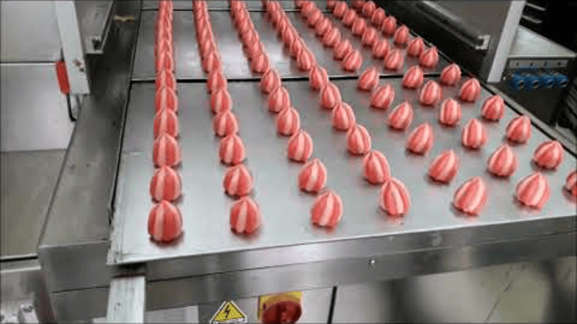Rows of red and white striped candies or confections are being formed and aligned on a metal conveyor belt in a factory setting.