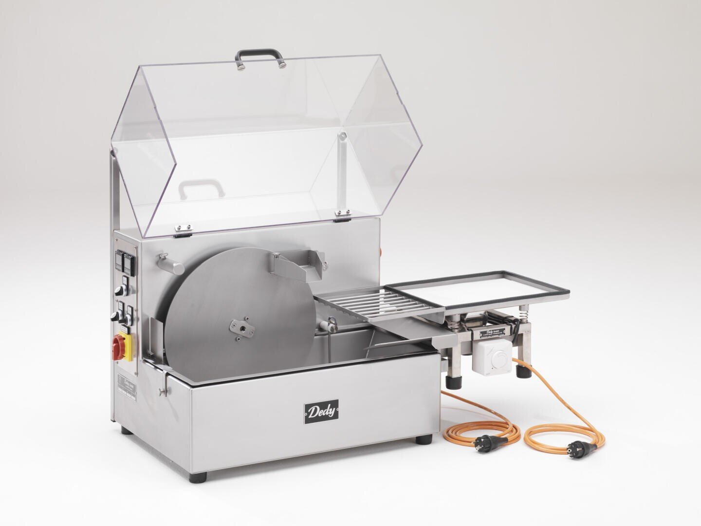 Eine industrielle Aufschnittmaschine aus rostfreiem Stahl mit einer Sicherheitsabdeckung aus durchsichtigem Kunststoff, einer runden Klinge, einem Bedienfeld und einem beigefügten Tablett auf einem weißen Hintergrund. Zwei orangefarbene Stromkabel sind daneben aufgewickelt.
