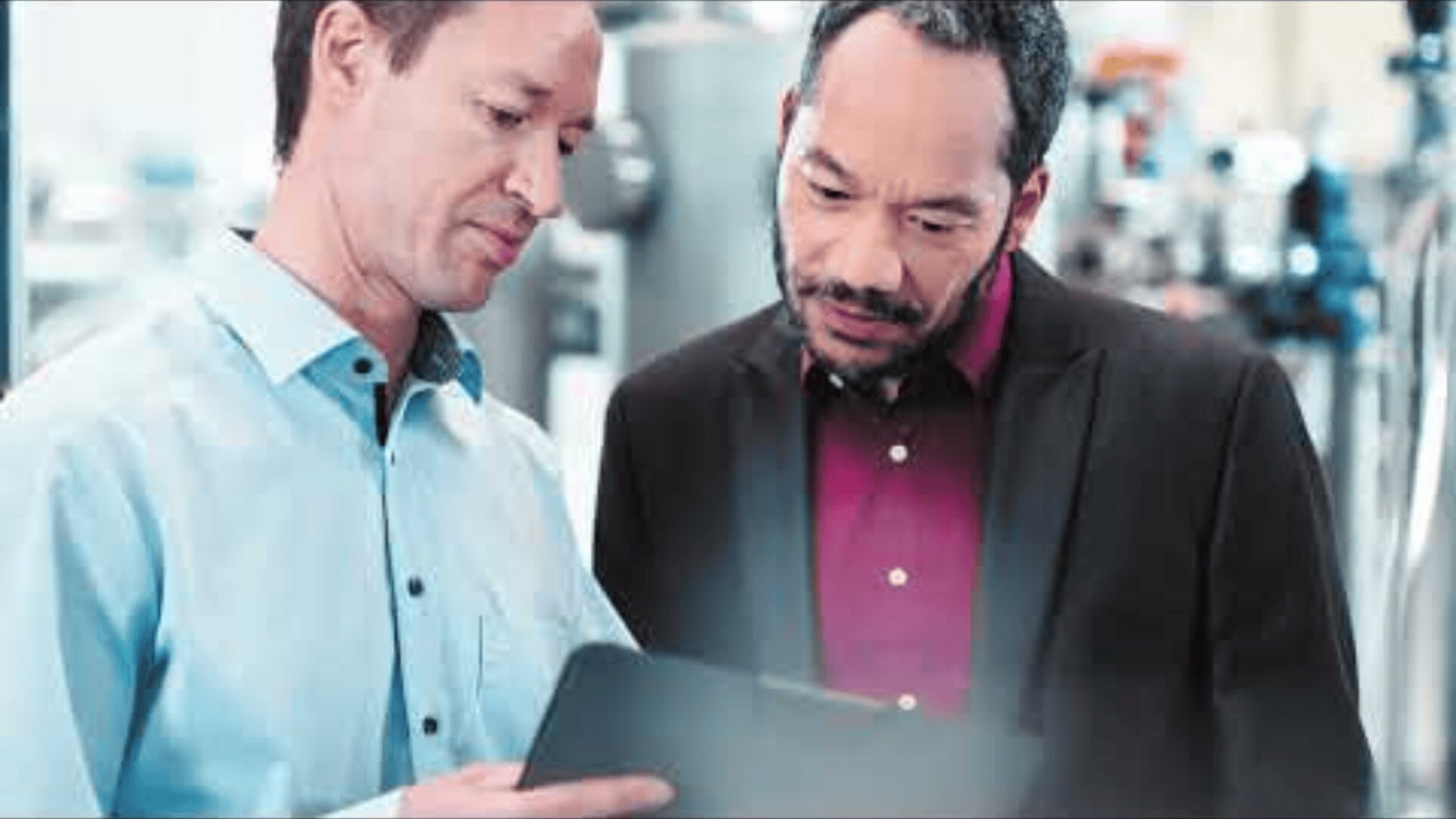 Zwei Männer stehen in einem Haus und schauen gemeinsam auf ein Tablet; einer trägt ein hellblaues Hemd, der andere einen dunklen Blazer mit rotem Hemd. Sie scheinen Informationen zu besprechen oder zu prüfen.