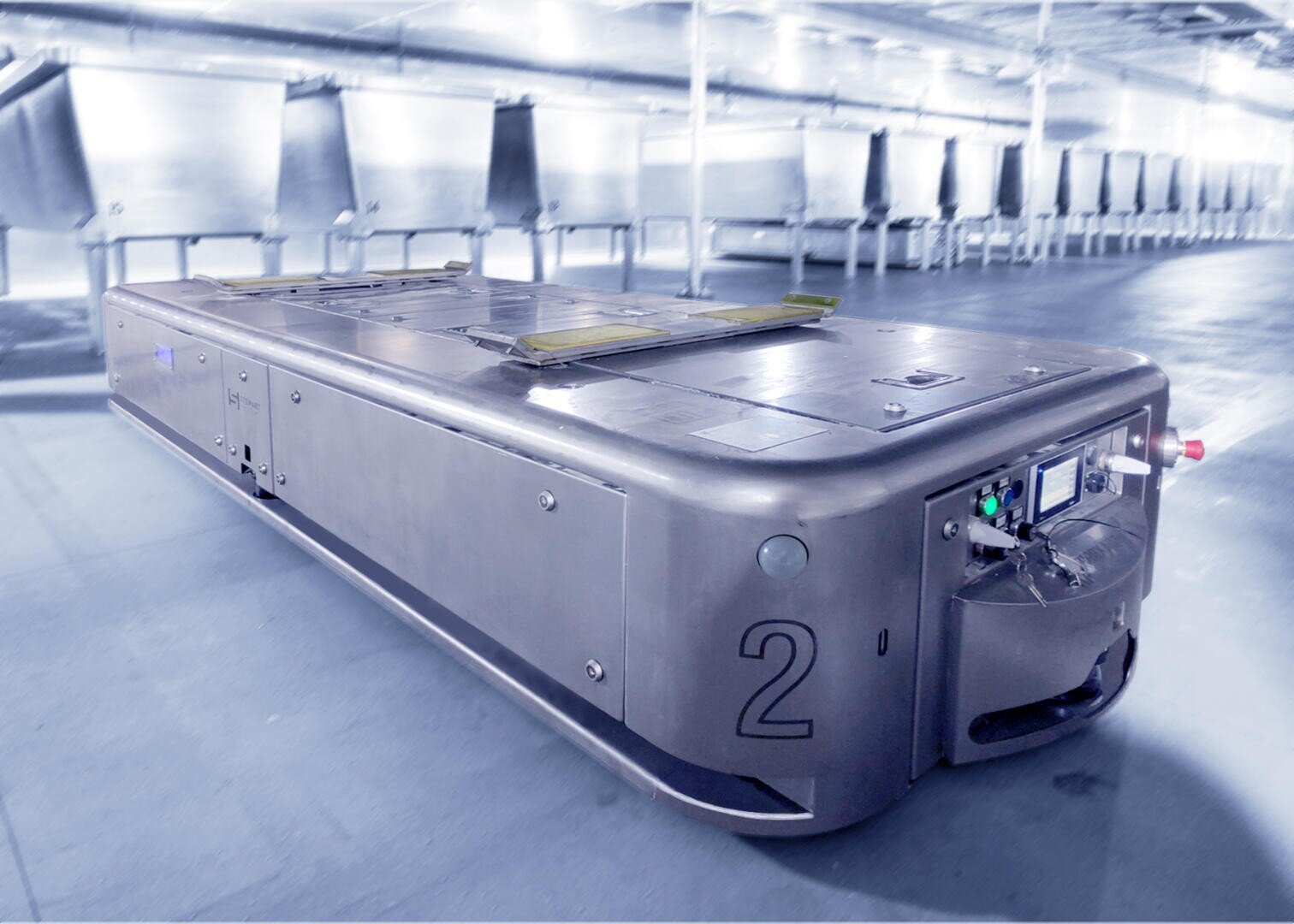 A large, rectangular automated guided vehicle marked with the number 2 is on the floor of an industrial facility, with empty metal racks or conveyor chutes in the background.