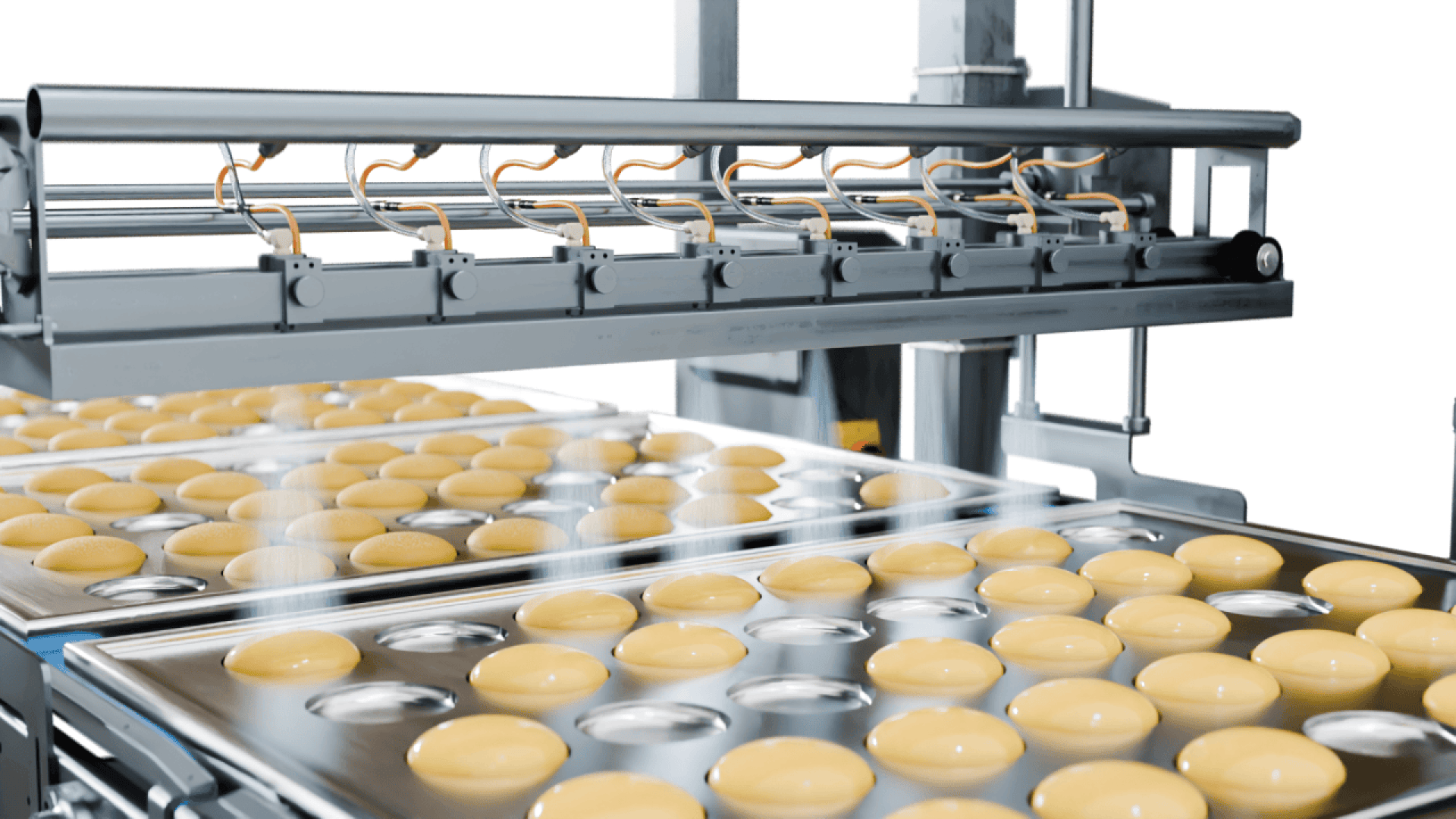 Reihen runder Teigstücke auf Metallblechen, die in einer Fabrik unter einer mechanisierten Back- oder Glasurmaschine hindurchlaufen, veranschaulichen einen industriellen Lebensmittelproduktionsprozess.