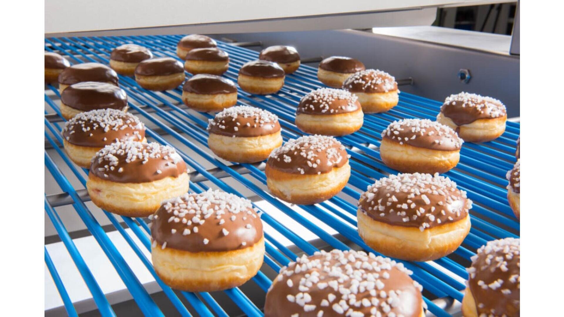 Reihen von Donuts mit Schokoladenglasur und weißen Streuseln bewegen sich in einer Fabrik auf einem blauen Förderband und deuten auf einen automatisierten Donut-Produktionsprozess hin.