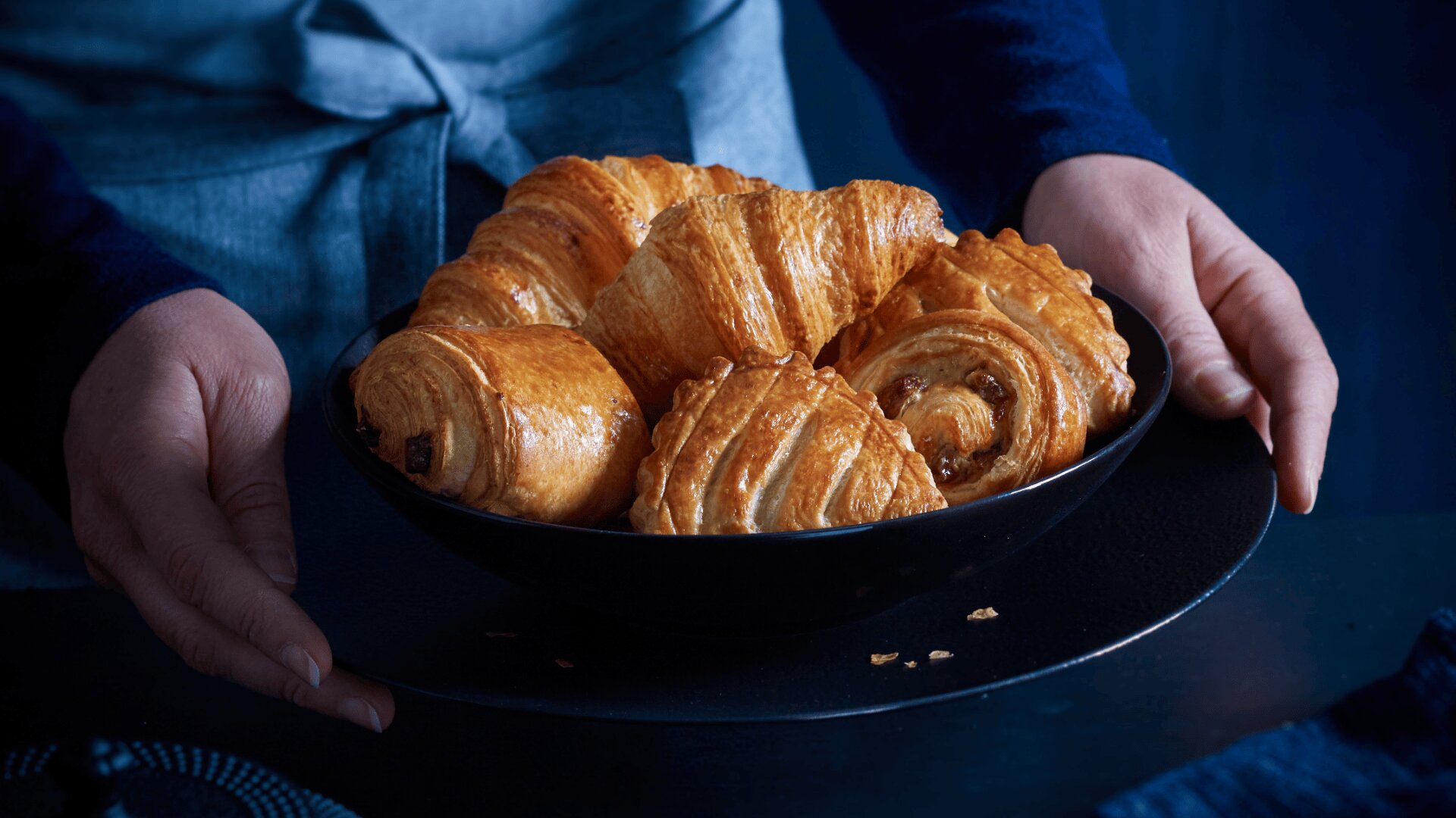 Eine Person hält einen schwarzen Teller mit verschiedenen goldbraunen Backwaren, darunter Croissants und Pain au chocolat, vor einem dunkelblauen Hintergrund.