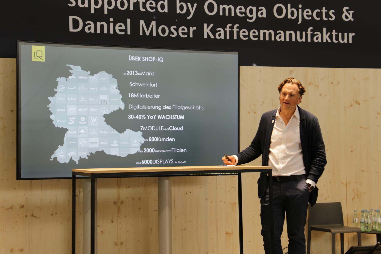 Ein Mann im Anzug steht neben einem Bildschirm, auf dem eine Präsentation über SHOP-IQ in deutscher Sprache läuft. Zu sehen sind eine Deutschlandkarte und Statistiken zu Unternehmenswachstum und Digitalisierung. Im Hintergrund sind eine Holzwand und Logos zu sehen.